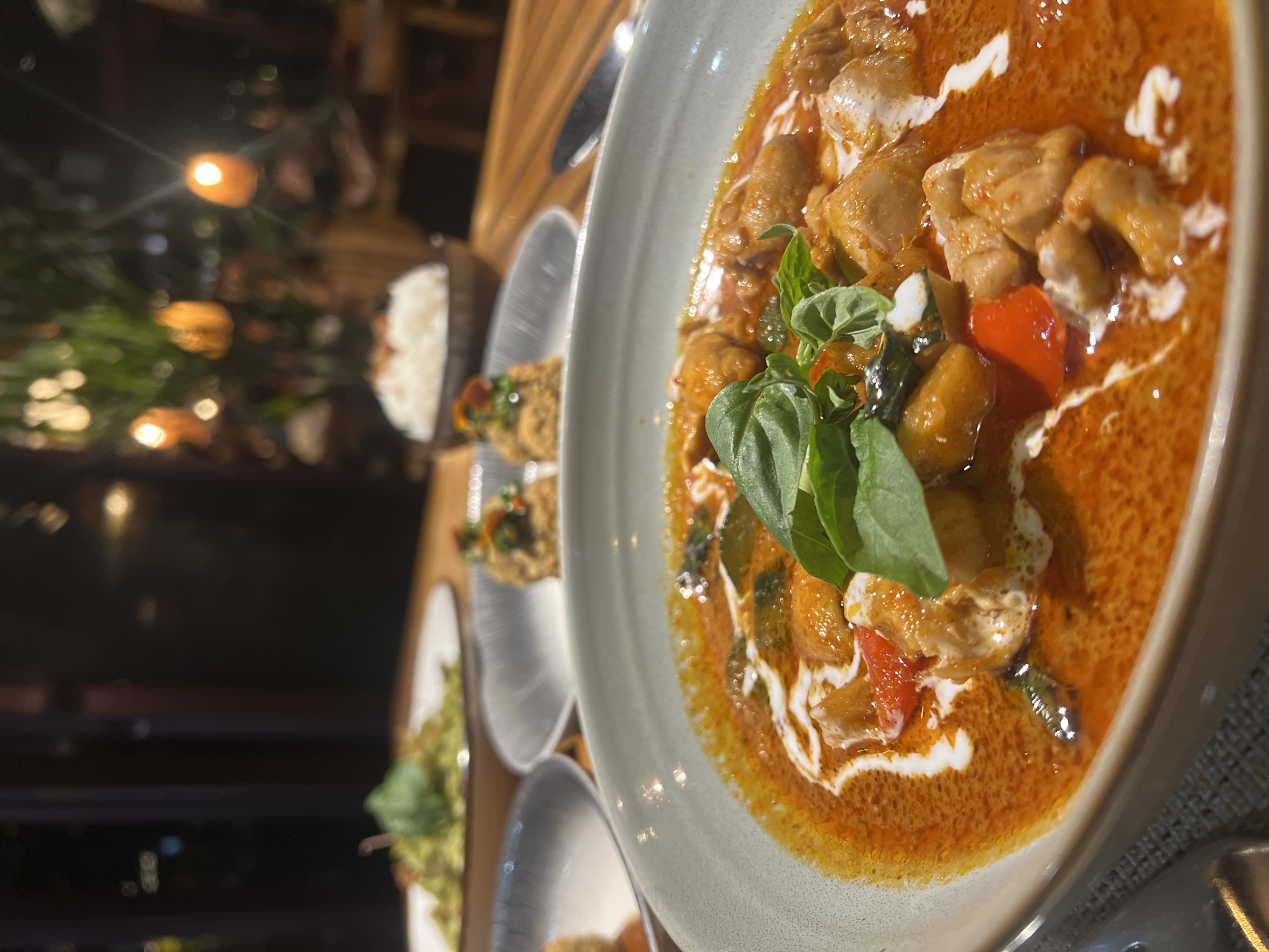 A close-up shot shows a table setting, likely in a restaurant or dining establishment in North Kuta, Indonesia. The main subject is a bowl of red curry with visible pieces of chicken, red peppers, and fresh basil leaves on top, finished with swirls of cream. Other dishes are visible in the background on the table, including a bowl of white rice and what appear to be other appetizers or side dishes. The lighting suggests it's evening or a low-lit interior with blurred lights in the background.