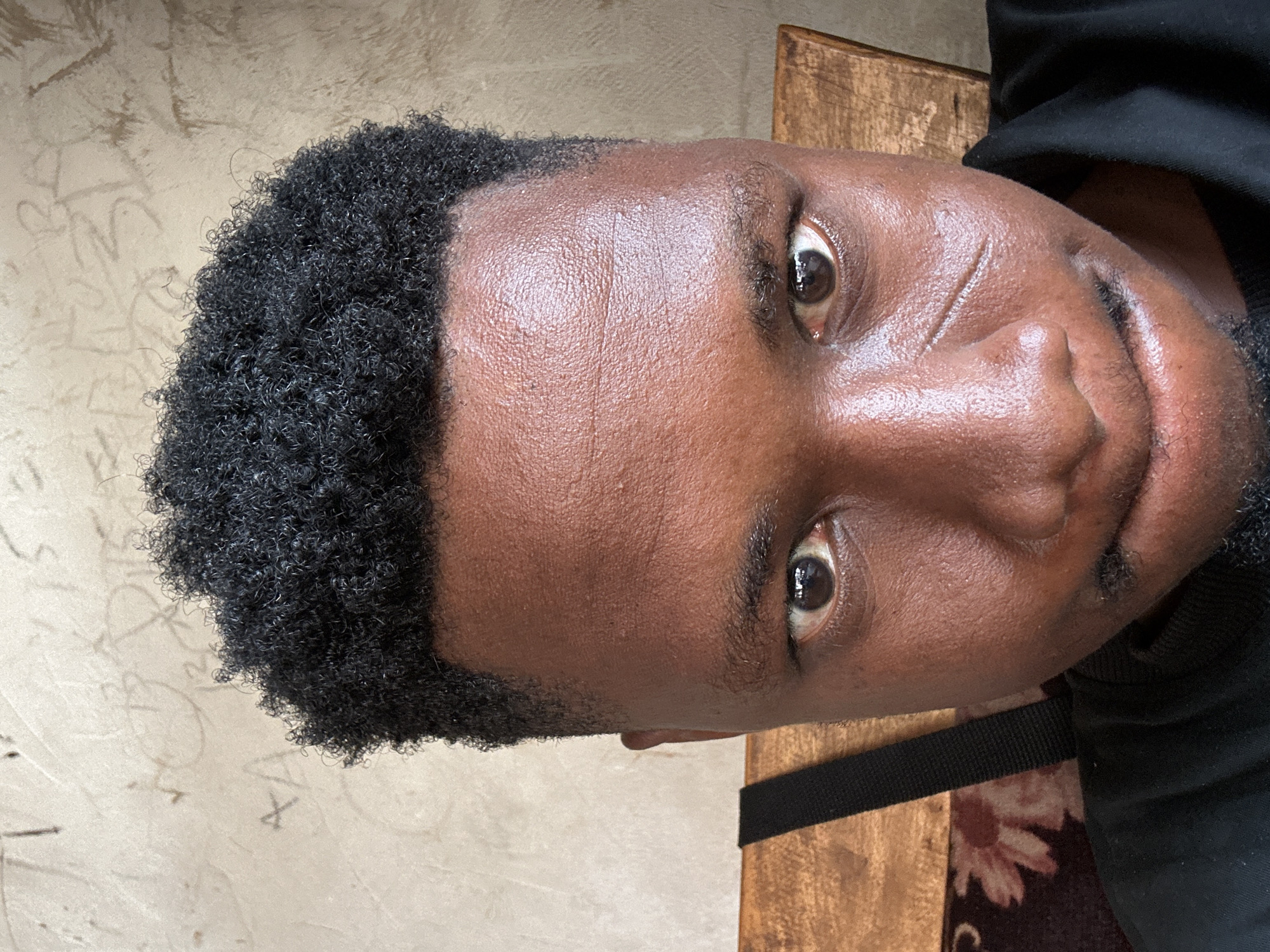 The image is a close-up, head-and-shoulders portrait of a young Black male taken indoors, likely in Mararaba, Nigeria. He has dark, curly hair, a dark complexion, and is looking directly at the camera. A noticeable scar is present on his right cheek, below the eye. He is wearing a black shirt. Behind him, a textured, light-colored wall with faint markings or scribbles is visible. To his left, a portion of a wooden chair and a dark strap, possibly from a bag, are visible. The lighting suggests an indoor setting, possibly with natural light from the front.