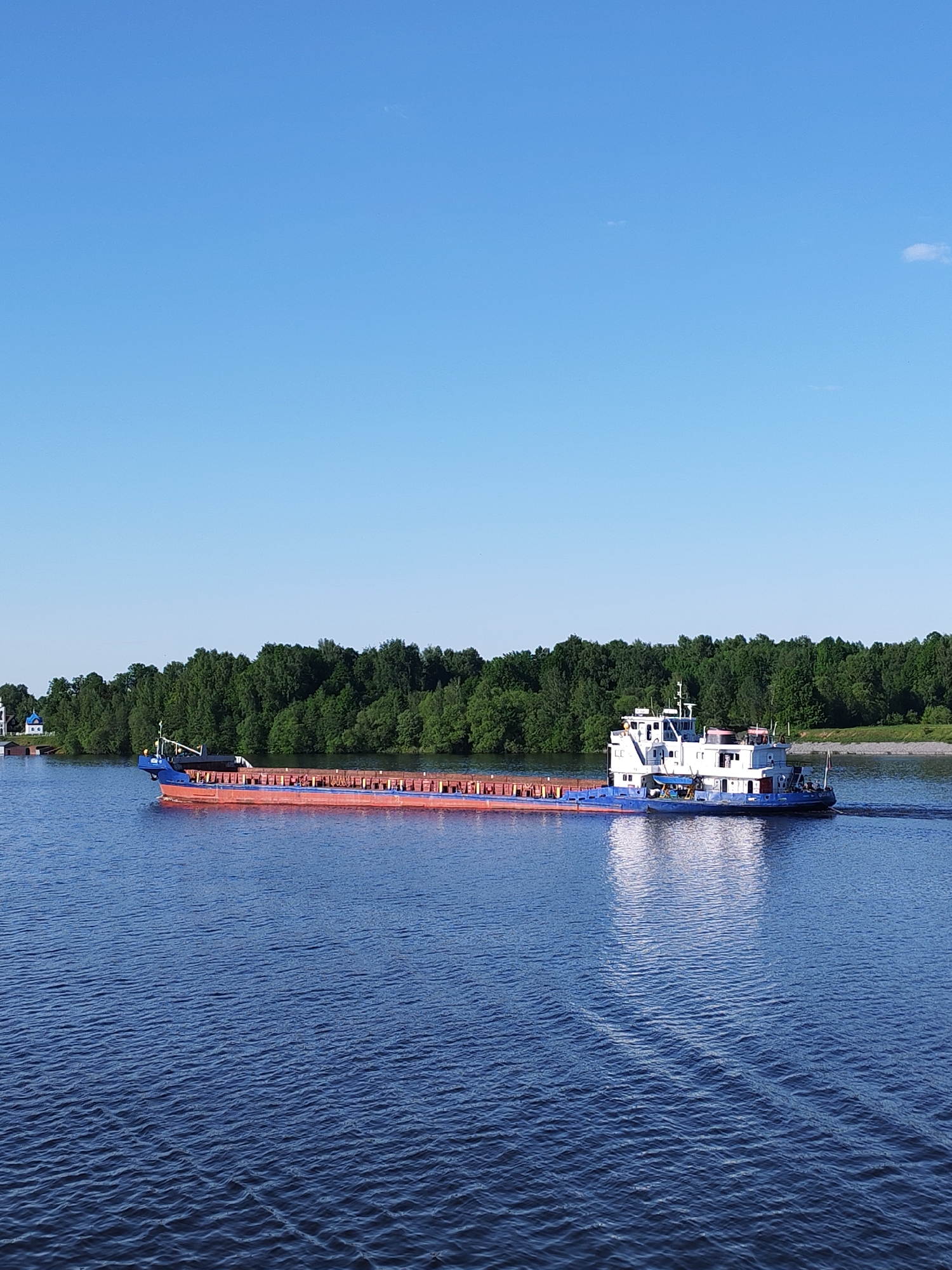 A long, flat-bottomed barge, loaded with cargo, moves across a body of water. The barge is blue and rust-colored with a white superstructure at the stern. It leaves a wake behind it. The water is deep blue with small waves. In the background, a line of dense green trees lines the far shore. A small building with a blue roof is visible on the left side of the frame, partially obscured by trees. The sky is clear and blue, with a very faint cloud in the upper right corner. The location and city are unknown.