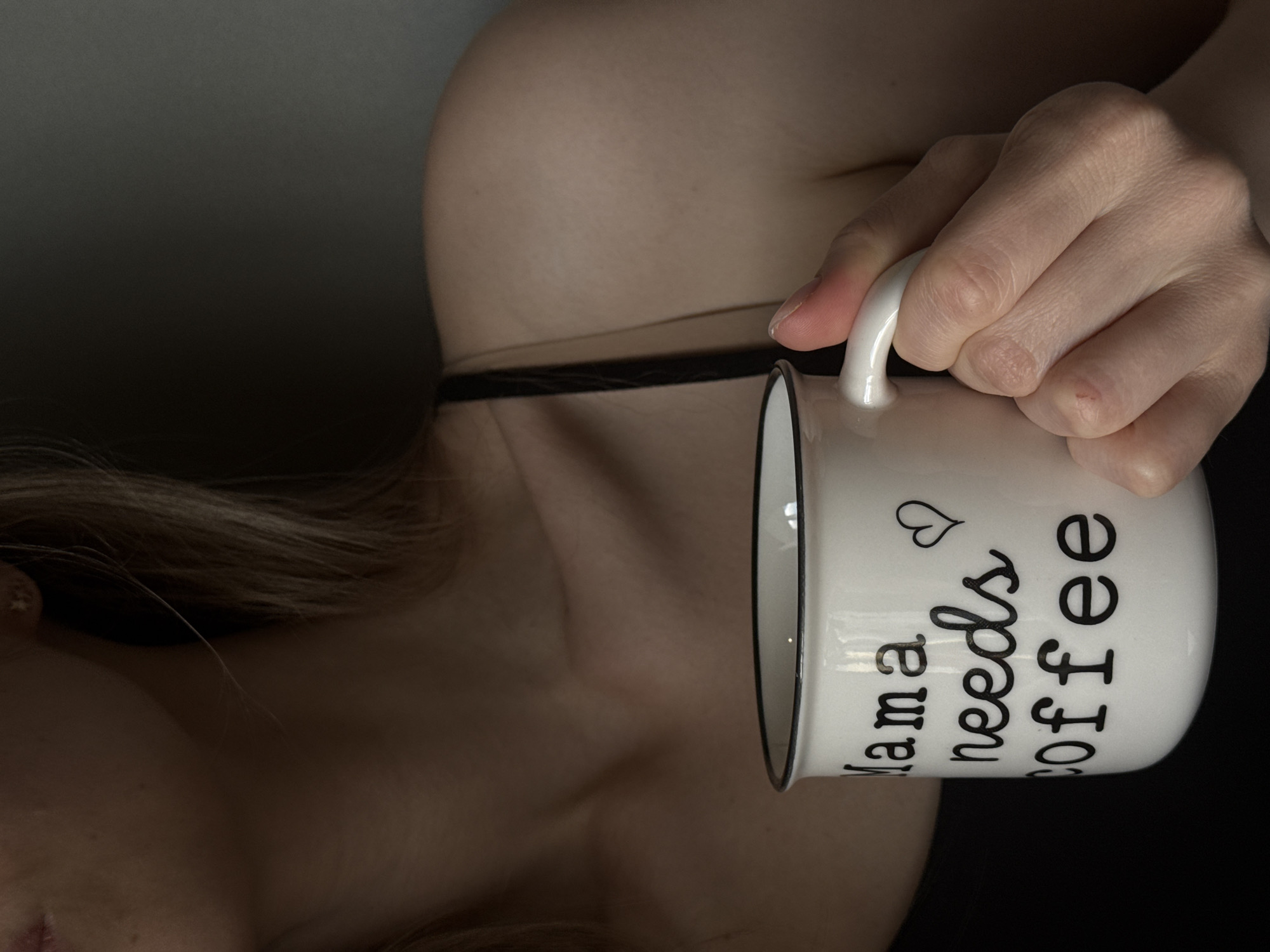 This close-up image, taken in Chaikovsky, Russia, features a person holding a white ceramic coffee mug. The photo is composed from an angled perspective, showing a bare shoulder, upper chest, and part of an arm. A thin black strap, possibly from an undergarment, is visible across the chest.
The person's hand is holding the mug by its handle, with the fingers wrapped around the cup. The mug has a black rim and handle and displays text in black script: "Mama needs coffee", accompanied by a small heart symbol above the word "needs". The background is dark and out of focus, drawing attention to the person and the mug. The lighting is soft, creating subtle contrasts on the skin. The overall atmosphere is intimate and suggests a quiet, personal moment, possibly in a relaxed indoor setting.