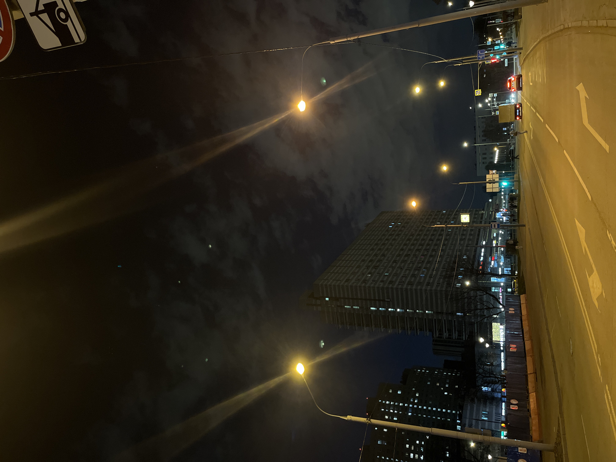 The image depicts a street scene in Moscow, Russia, at night. The road is illuminated by streetlights, with white directional arrows painted on the asphalt. Several cars are visible on the street. A tall, modern building with many illuminated windows dominates the background, along with another large, lit building. Streetlights are strung along the road. The sky is dark and cloudy. The overall impression is that of a city street at night.