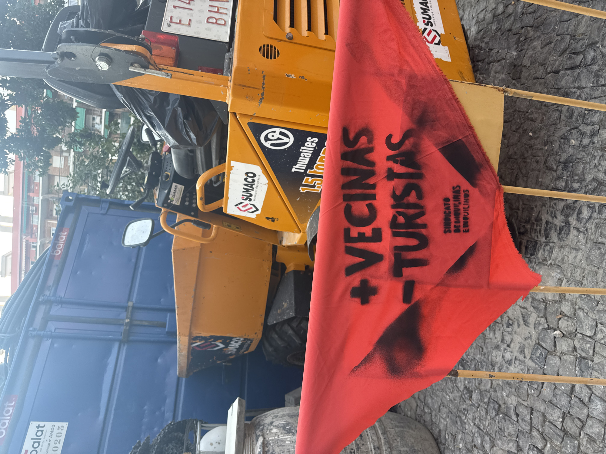 The image shows a red flag with black spray-painted text protesting against "Vecinas Turistas," which translates to "Tourist Neighbors" or "Neighbor Tourists." The flag is draped over yellow construction equipment, possibly a roller or compactor, manufactured by Thwaites and possibly branded SUMACO. The text on the flag also includes "SINDICATO DE INQUILINAS E INQUILINOS," suggesting a union or collective of renters.
In the background, a blue truck with the branding "Balat" is visible, along with trees and buildings, hinting at an urban setting. The ground appears to be paved with cobblestones. The overall scene suggests a protest or demonstration, likely related to housing issues and the impact of tourism on local communities in Madrid, Spain. The lighting and shadows do not provide clear indicators of the time of day or specific weather conditions, but the scene is likely outdoors. The presence of construction equipment could indicate ongoing development or maintenance in the area, which might be a point of contention for the protesters.