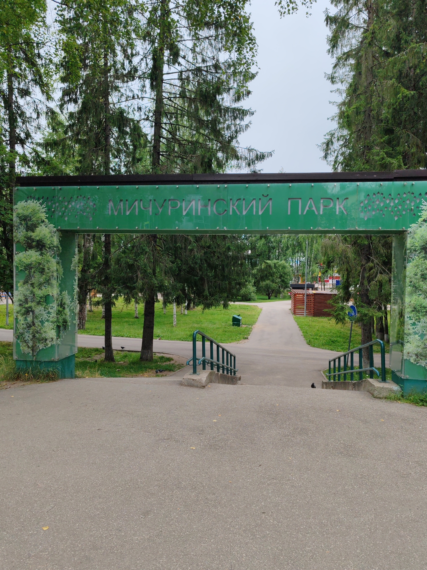 The image shows the entrance to Michurinsky Park in Syktyvkar, Russia. A green archway with decorative patterns and the park's name in Russian ("МИЧУРИНСКИЙ ПАРК") spans across a paved pathway. The archway is flanked by tall evergreen trees and features stylized depictions of trees on its pillars. Beyond the archway, a pathway leads into the park, with a wooden structure and playground equipment visible in the distance. The ground is paved with asphalt, and there are some green spaces with trees and grass. Two birds are visible on the path. The sky is overcast.
