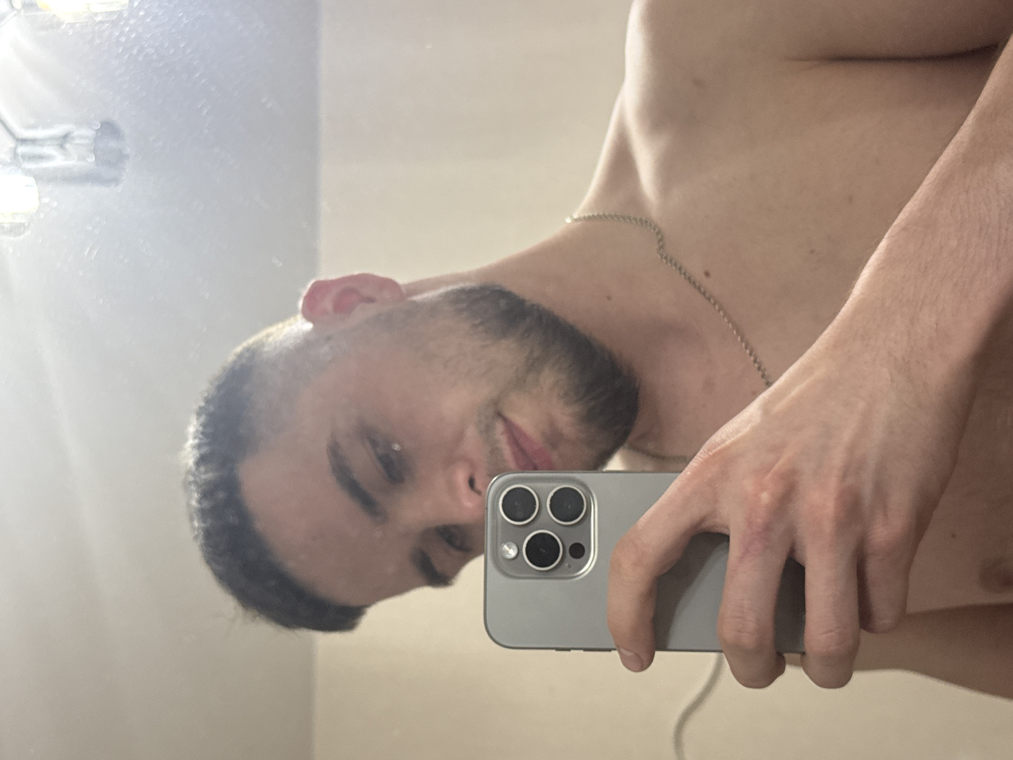 The image is a mirror selfie featuring a shirtless young man, likely taken indoors in Perm, Russia. The man is reflected upside down and slightly angled, holding a gray/silver smartphone with his right hand, positioned in front of his lower face.
**Subjects:** The primary subject is a male with light skin, short dark hair, and a trimmed beard. He is wearing a thin, silver-colored chain necklace around his neck. His visible expression is neutral to slightly smiling. The smartphone, likely an iPhone Pro model, displays a prominent triple-lens camera system with a flash module. Parts of a light fixture or lamp are visible in the upper left corner of the reflection.
**Activity:** The man is actively taking a mirror selfie, looking at his reflection and the phone's camera.
**Notable Details:** The background reflected in the mirror appears to be an indoor setting, characterized by light-colored walls, possibly a bathroom or bedroom. There's a prominent bright light source reflected in the upper left. The man's body shows some muscular definition in the shoulders and chest area. His hand holding the phone has visible veins. The mirror surface exhibits some dust or specks.
**Visible Text:** There is no visible text in the image.