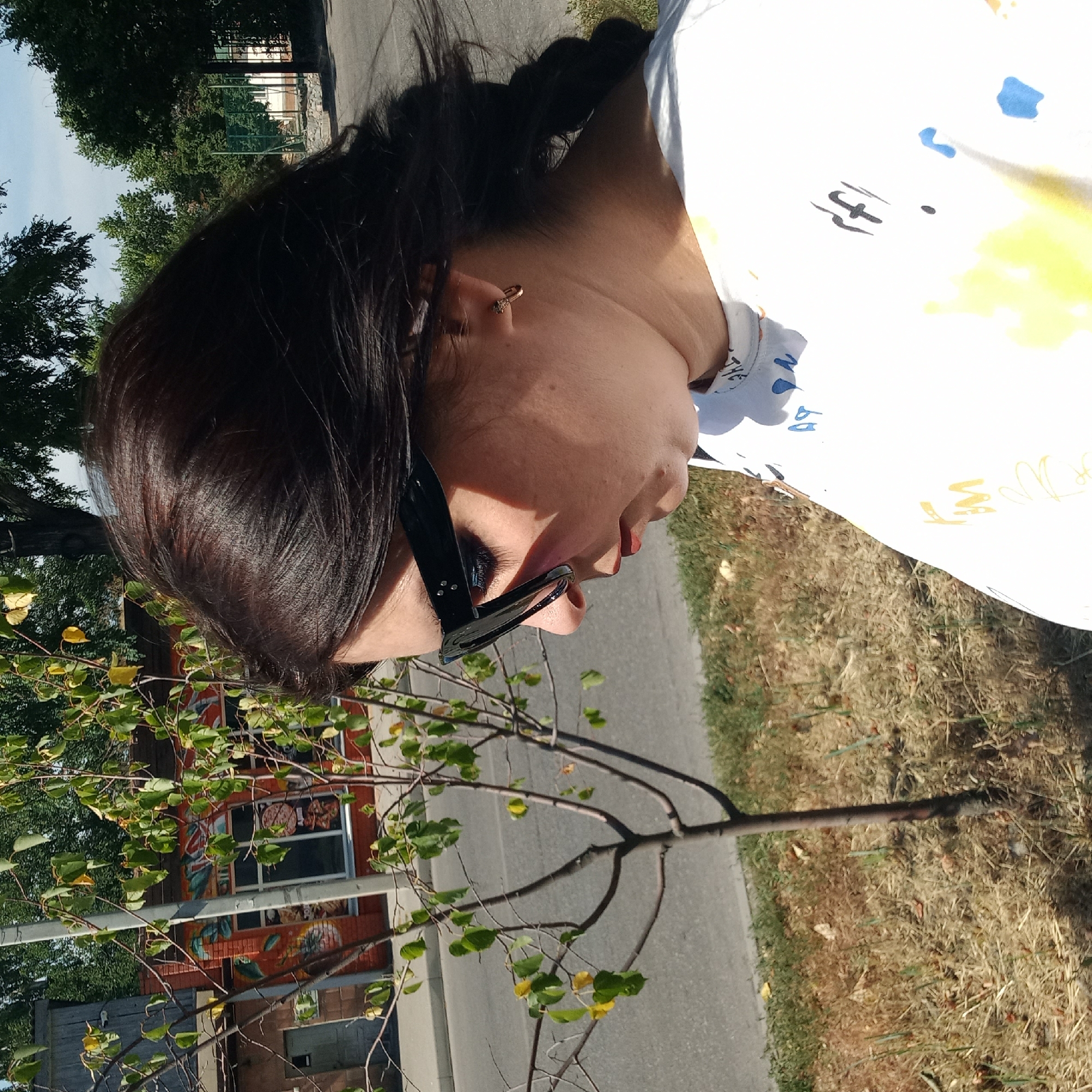 A medium shot captures a woman from the chest up, her body angled to the right and her head turned to look down. She has dark hair styled in a braid and wears black-rimmed sunglasses and a white t-shirt with abstract colorful markings. In the background, a tree with sparse green and yellow leaves stands near a street with a sidewalk. Further back, a brick building with windows and graffiti-style art is visible. The scene is set outdoors under a clear blue sky, likely on a sunny day. The presence of a basketball court in the distance, along with residential-style buildings, suggests a neighborhood or park area within the city of Kropyvnytskyi, Ukraine.