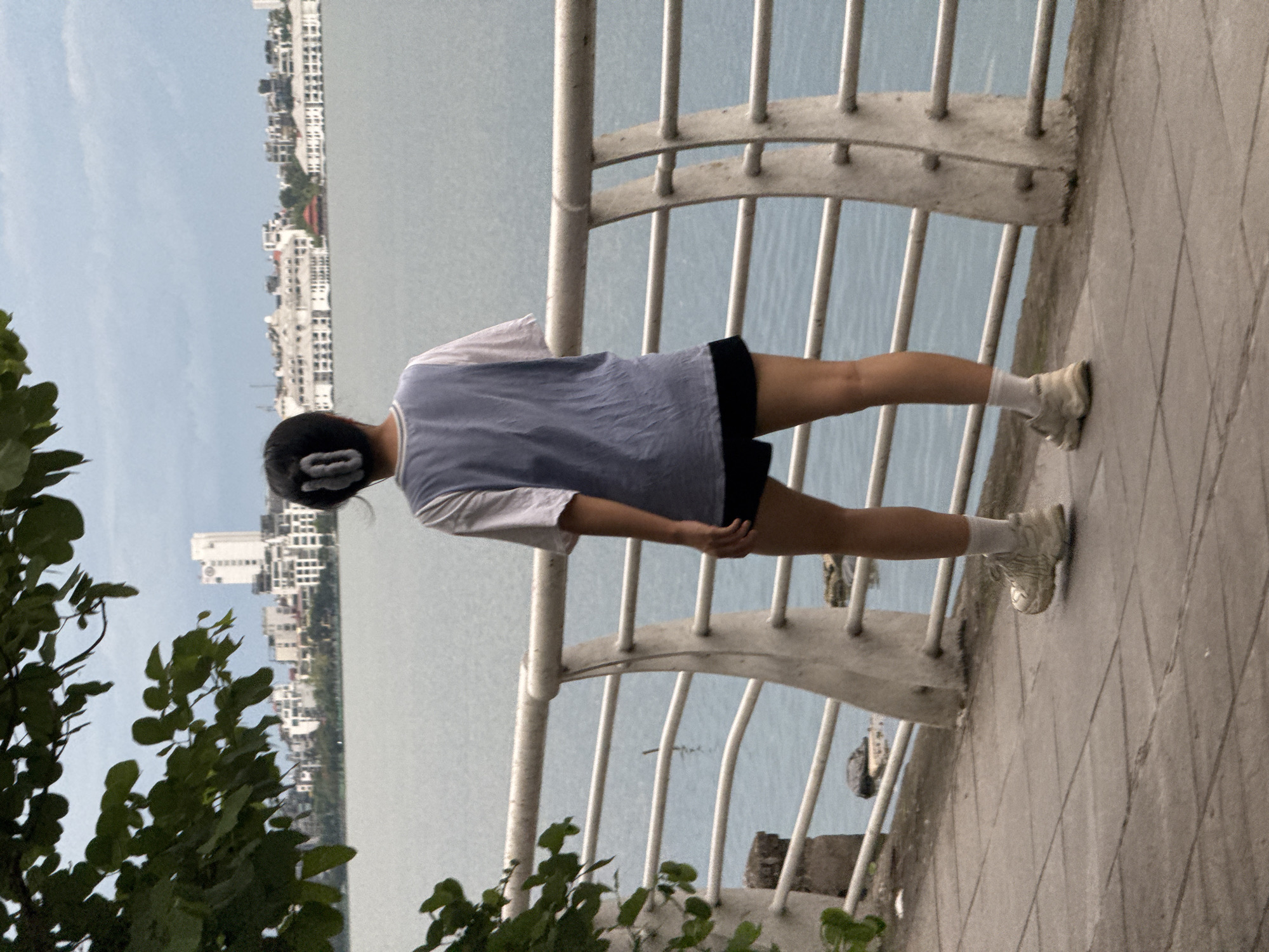An individual stands on a tiled walkway facing away from the camera, looking out over a body of water towards a city skyline. The person is wearing a light blue t-shirt over a white t-shirt, black shorts, white socks, and light-colored sneakers. Their dark hair is pulled back with a white hair clip. A white railing separates the walkway from the water. In the background, a dense cluster of white buildings, some with multiple stories, forms the cityscape. The sky is overcast with a soft, diffused light. The location is likely the West Lake area in Hanoi, Vietnam, known for its extensive waterfront and urban development. Lush green leaves from a tree are visible in the foreground, framing the left side of the image. The water appears calm, with ripples suggesting a gentle breeze.