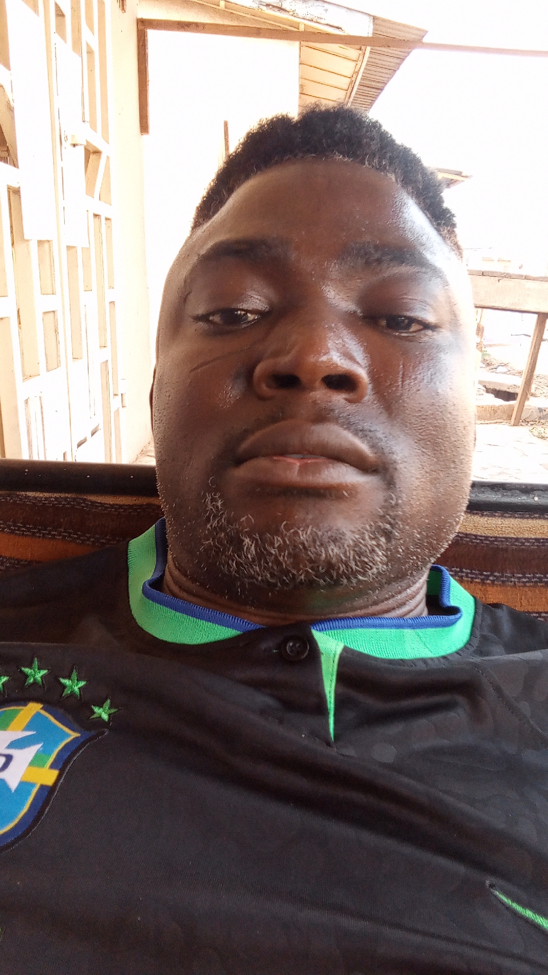 A close-up photograph depicts a dark-skinned man with short, dark hair and a graying beard. He has what appears to be a scar on his left temple and minor marks on his face. He is wearing a dark-colored shirt with a green and blue collar. The shirt has a logo on the left side with green stars and a blue and yellow emblem. The background includes a building structure, possibly a veranda or porch. The image was taken during daylight, as indicated by the lighting. It's impossible to confirm the location specifically as Sabon Tasha, Nigeria without further metadata or contextual clues within the image itself. The identification of the city relies on information outside of the image.