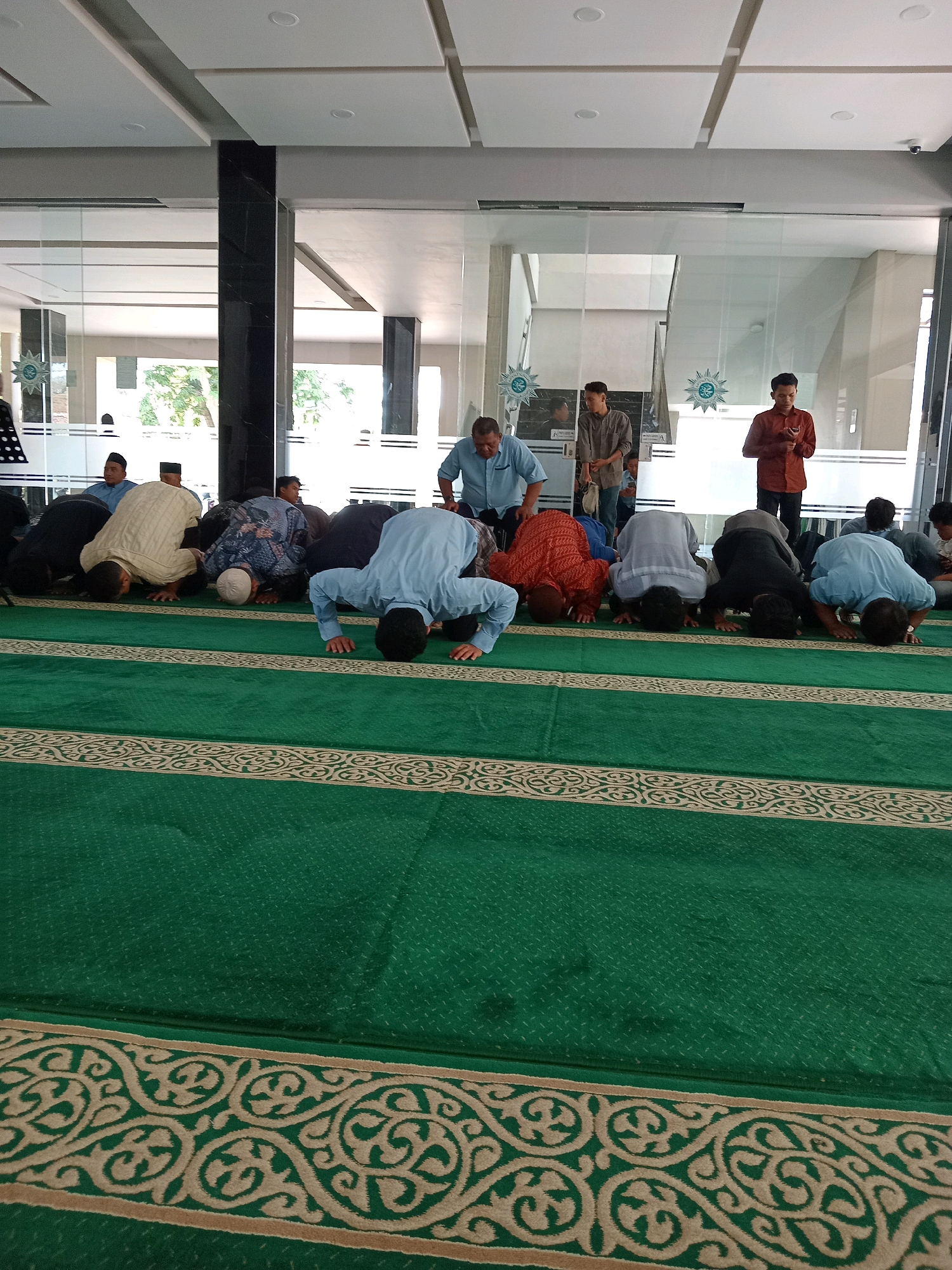 The photograph captures a group of men performing the Sujud (prostration) in a prayer hall. They are arranged in rows on a green carpet with decorative borders, indicating a place of worship. The architecture suggests a modern building with large glass windows and a tiled ceiling. In the background, several other individuals are visible, some standing and observing. One man in a reddish-brown shirt appears to be looking at his phone. The lighting is bright, likely from the overhead fixtures and natural light filtering through the windows. The overall scene depicts a religious gathering in a well-maintained indoor space. The image is taken from a slightly elevated perspective, looking down at the praying individuals.