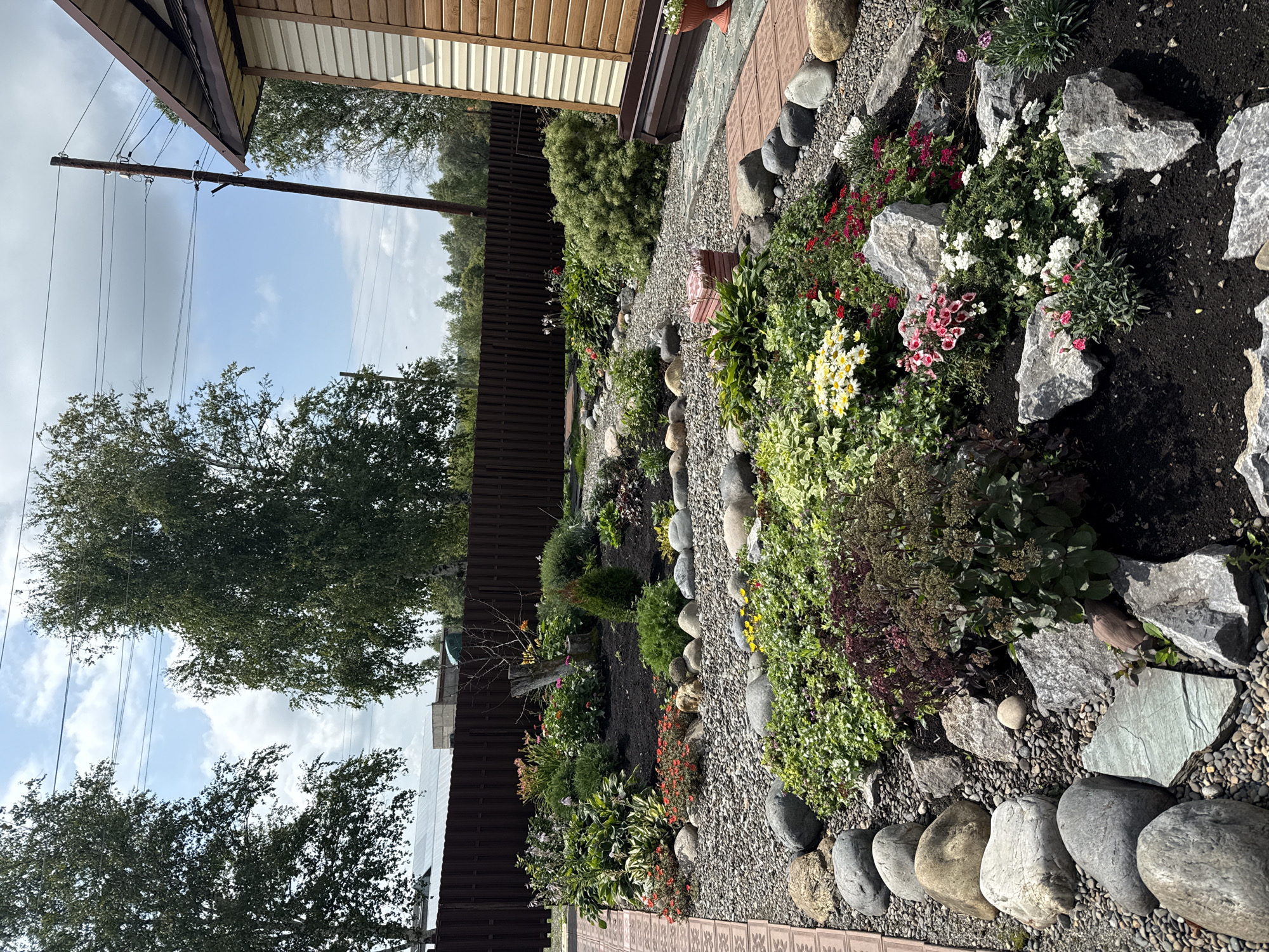 The image depicts a well-maintained garden area, likely a residential property, in the village of Krasnogorskoye, Russia. The scene features a variety of plants and flowers, including hostas, sedums, daisies, and smaller flowering plants, arranged in raised beds and along pathways. The garden is bordered by large, rounded river stones and smaller gravel. A brown wooden fence encloses the property in the background. To the right, a section of a wooden-clad building with a brown roof is visible. Overhead, power lines stretch across a partly cloudy sky. The overall impression is one of a peaceful, natural outdoor space.
