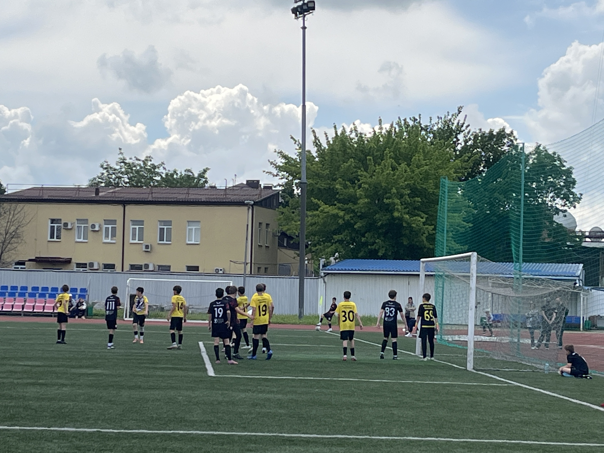The image shows a soccer game taking place on a green field. Players in yellow and black uniforms are on the field. Other individuals are seen standing near the goal. The sky is cloudy. In the background, there is a yellow building and trees. The city of Krasnodar, Russia cannot be determined from the available visual information.
