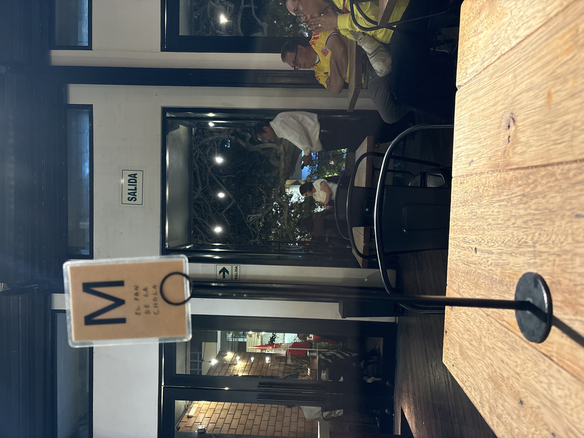 In San Isidro, Peru, a dimly lit restaurant patio features patrons seated at tables. A sign with a large "M" and Spanish text "EL PAN DE LA CHOLA" is prominently displayed in the foreground. To the right, a "SALIDA" (Exit) sign is visible on the wall. Several individuals, some wearing yellow and red, possibly representing the Colombian flag, are seen conversing or observing. The ambiance suggests a casual dining or gathering setting.