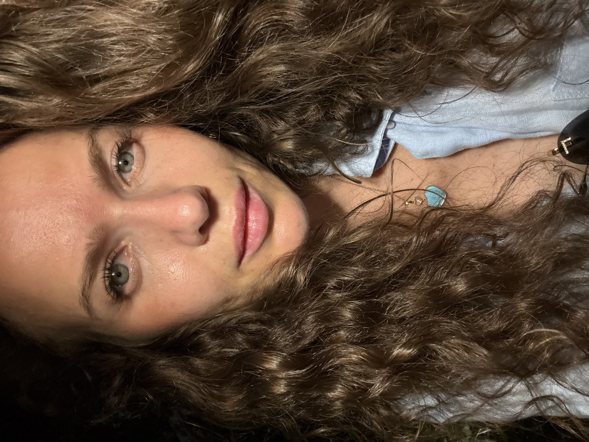 The image is a close-up portrait of a woman with long, curly brown hair and blue eyes. She is wearing a light blue collared shirt and a necklace with a turquoise heart-shaped pendant. Her skin appears slightly flushed. The background is dark and out of focus, with some subtle hints of greenery on the left side. There is no discernible indication of a specific location in Dubai, United Arab Emirates.