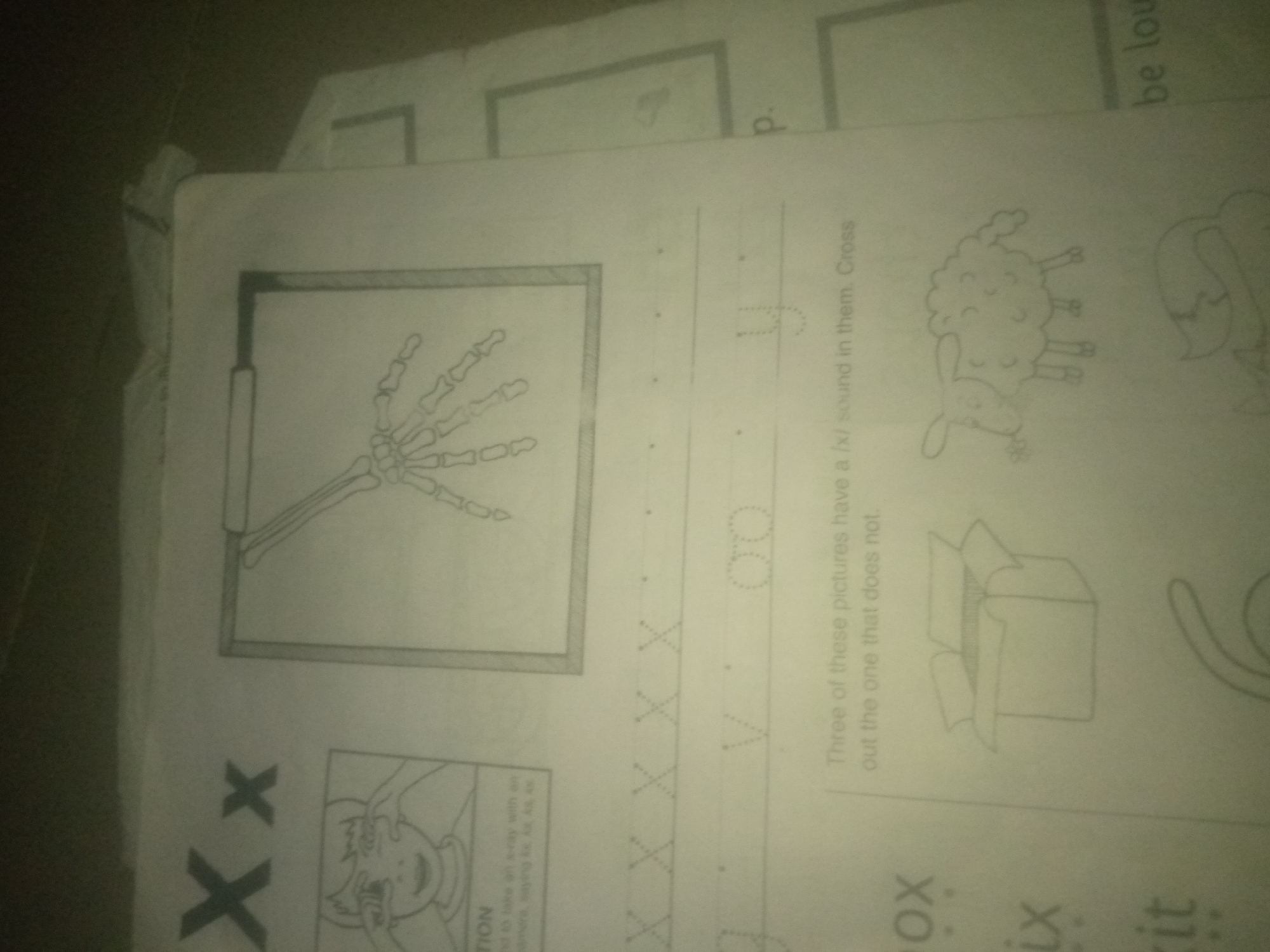 The image is an overhead shot of a children's worksheet with several drawings and some text. On the left side of the page is a detailed drawing of a human hand skeleton inside a rectangular frame. To the right of the skeleton, there are dotted lines forming the letter "X" several times, followed by a dotted outline of the letter "U" and then two dotted circles. Below these are the words "Three of these pictures have a /x/ sound in them. Cross out the one that does not." There are also small drawings of a sheep and an open box. In the bottom left corner, the letter "X" is written twice in large print. Below that, there is a partially visible drawing of a child's face. Further to the right, at the bottom of the page, are the words "ox," "ix," and "it" with dotted lines. A partial drawing of what appears to be a fox tail is visible on the far right. The lighting in the image is dim and casts a shadow over the page, making some details difficult to discern. The overall context suggests an educational activity focused on letter recognition and phonics. There is no indication of the city of Jauro Yinu, Nigeria within the image.