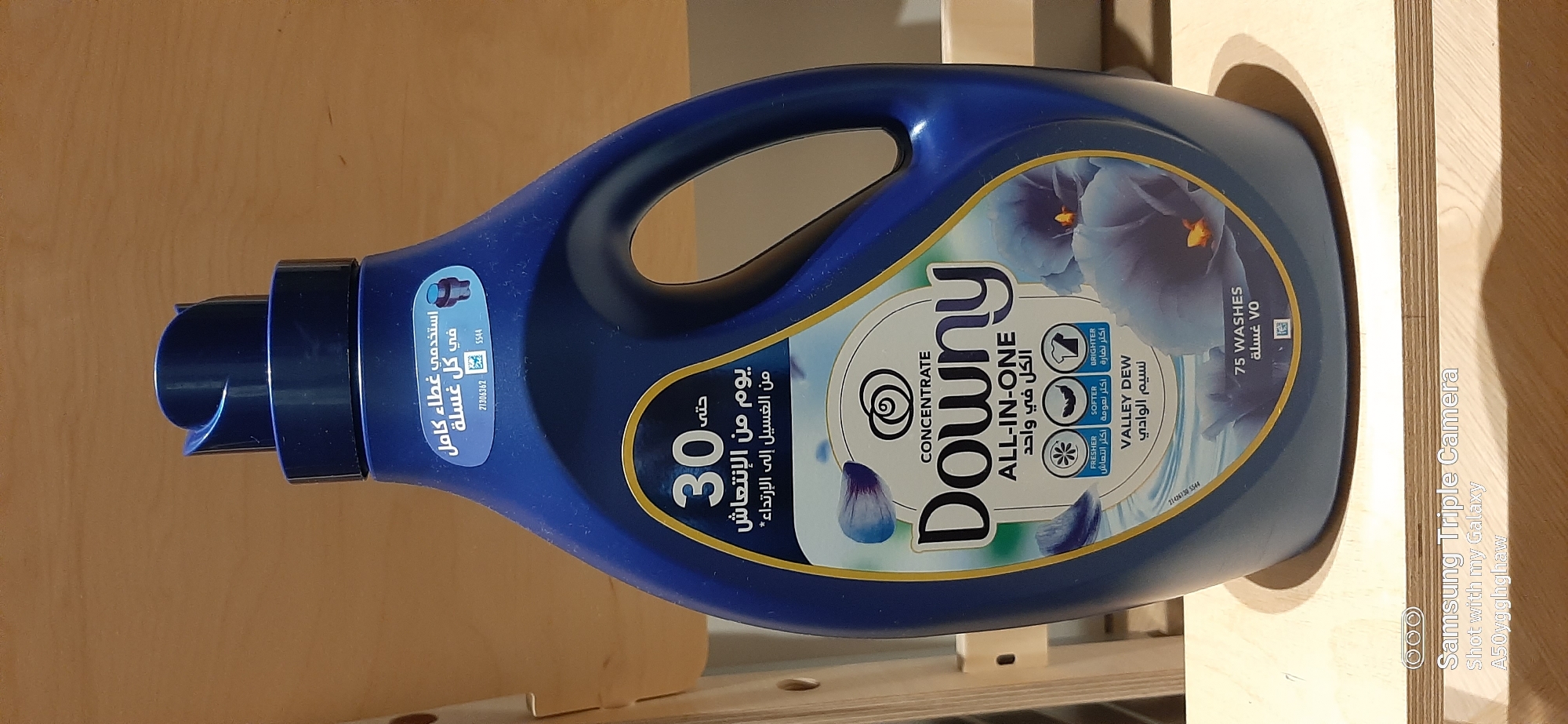 A blue bottle of Downy fabric softener is displayed against a plain wooden background. The bottle features a prominent yellow and white label with Arabic text and graphics of blue flowers. The label indicates that the product provides 30 days of freshness and is "All-In-One," with icons representing freshness, softness, and brightness. The scent is identified as "Valley Dew." The bottle has a dark blue cap and a built-in handle. The overall setting suggests an indoor environment, possibly a home or retail display, given the lighting and the wooden surfaces. There are no people visible in the image. The image is tagged with "Samsung Triple Camera" and "Shot with my Galaxy A50yggghaw," indicating it was taken with a Samsung phone.