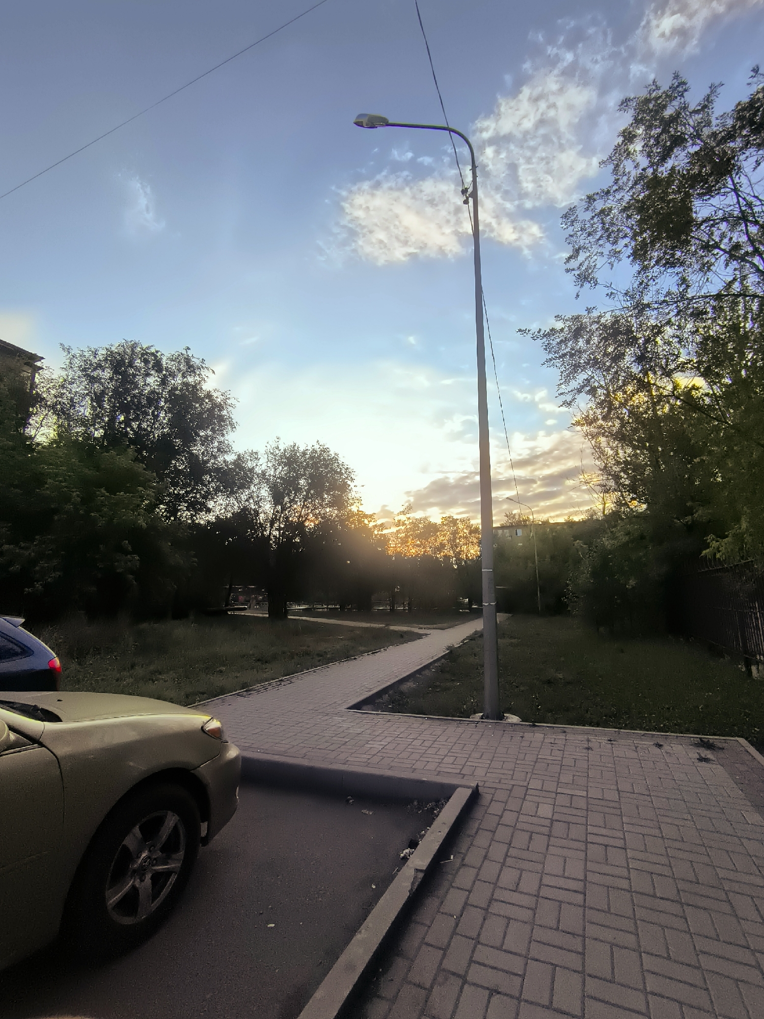 An eye-level, medium shot captures a paved walkway curving through a grassy area with trees lining the path and a street lamp positioned centrally. In the foreground, the hood and front wheel of a light-colored car are visible, parked on asphalt. A dark car is partially seen on the left. The sky is a mixture of blue and white clouds, with the sun setting in the distance, casting a warm glow through the trees. The scene appears to be in a residential area.