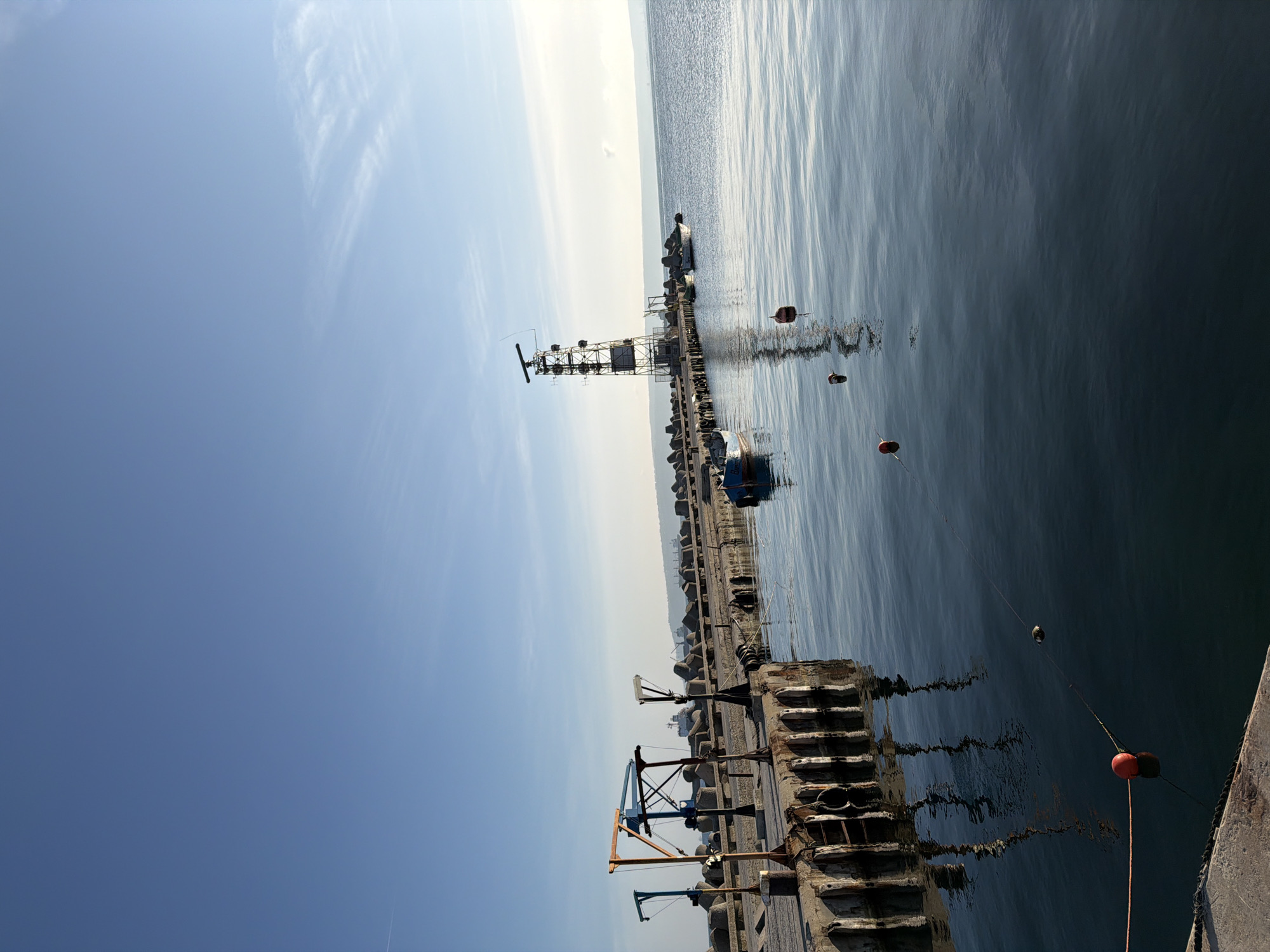 The image depicts a port scene in Varna, Bulgaria, under a clear blue sky with minimal cirrus clouds. A long concrete pier extends from the left foreground into the background, supporting various rusty metal structures resembling small davits or cranes, and numerous mooring bollards. The pier's edge reveals vertical concrete supports with visible wear. Adjacent to the pier's exterior, large grey concrete breakwater blocks are arranged.
In the midground, a blue fishing boat is moored to the pier on the left. Further down the pier, a tall, grey metal lattice tower stands prominently, equipped with a rotating radar dish and antennae at its summit. Several other small boats are visible moored near the tower.
The water, calm and dark blue, reflects the pier structures and the sky. Two prominent red buoys and one smaller dark buoy are visible floating on the water in the foreground and midground, attached by lines. The sea stretches to the horizon on the right, with distant land formations and a faint outline of a cargo ship visible. No individuals are present in the scene.