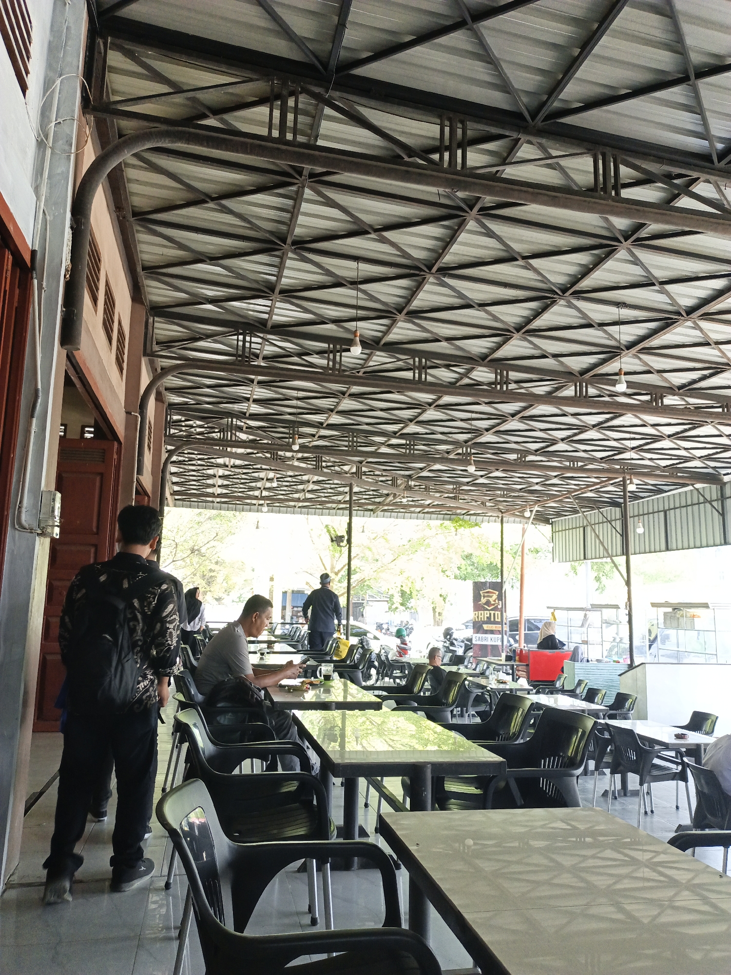 This image depicts a casual, open-air cafe or restaurant in Lhokseumawe, Indonesia, during the daytime. The setting features a large, industrial-style metal truss roof covered with corrugated metal sheets, under which numerous rectangular tables and dark plastic chairs are arranged. Several bare light bulbs hang from the roof structure.
In the foreground, a man wearing a patterned long-sleeved shirt and a black backpack stands with his back to the viewer, facing deeper into the cafe. To his right, another man in a grey t-shirt is seated at a table, looking down at an object on the table, possibly a mobile phone. A mug or glass is also visible on his table. Other patrons are scattered throughout the cafe, some seated and some standing, with a person in a hijab visible further back.
The background shows more outdoor elements, including motorcycles parked, indicating a roadside or accessible location. A black banner with white text is clearly visible, displaying "RAPTO," "JEMAS MAN," and "SABRI KUPI." "Kupi" is an Acehnese word for coffee, suggesting this is a coffee shop.
The scene is brightly lit by natural daylight, casting shadows and indicating clear weather. The overall atmosphere appears relaxed and functional, typical of a casual eatery.