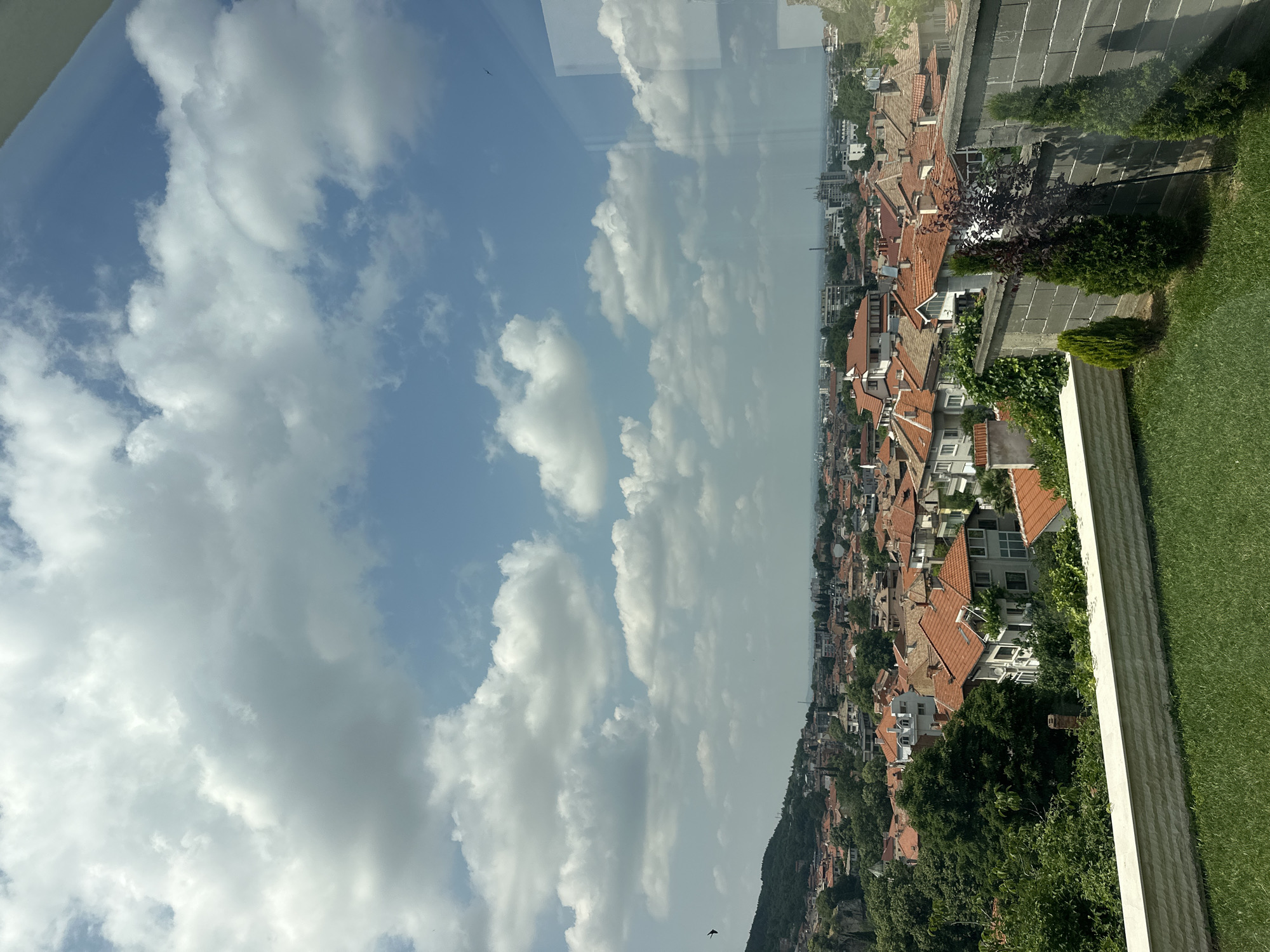 The image presents a panoramic view of Asenovgrad, Bulgaria, captured from an elevated perspective, likely through a window given the faint reflections visible. The scene is dominated by a sprawling urban landscape of residential buildings nestled against a backdrop of hills and a wide sky.
In the foreground, a neatly trimmed green lawn occupies the right side, bordered by a light-colored, textured wall running horizontally. Beyond this, the cityscape unfolds, characterized by numerous houses with distinctive orange or terracotta-tiled roofs. These buildings vary in size and style, with some appearing to be multi-story, and are interspersed with lush green trees and shrubs, suggesting well-integrated greenery within the urban fabric.
The city extends into the midground, with more buildings gradually fading into the distance. On the far left, a significant portion of a verdant, forested hill or mountain rises, adding a natural element to the urban sprawl. The horizon is somewhat hazy, suggesting either distance or atmospheric conditions like summer heat.
The sky is bright blue, adorned with scattered, fluffy white cumulus clouds, indicating fair weather. The lighting is strong and clear, consistent with daytime, probably late morning or early afternoon. No specific activities or people are visible, focusing instead on the architectural and natural landscape. There is no visible text in the image. The architectural style, particularly the tiled roofs, aligns with the typical appearance of towns in the Balkan region, confirming the Asenovgrad context.