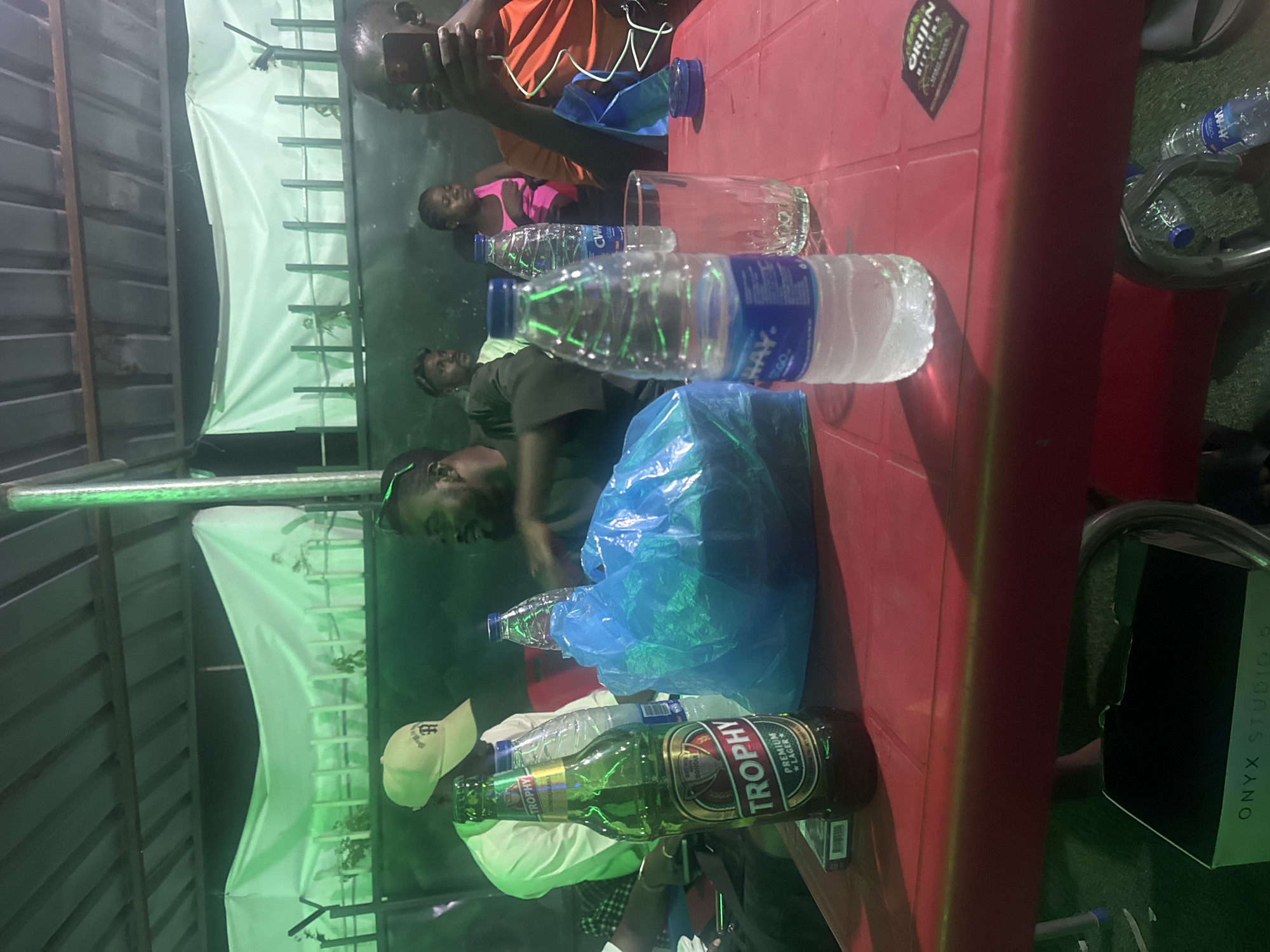 The image shows a gathering of people around a red table. On the table are multiple water bottles, a bottle of Trophy beer, and a drinking glass. The location is Karu, Nigeria, based on the beer brand "Trophy Lager." The scene appears to be outdoors, with a makeshift tent or awning visible. People are sitting and standing around the table, engaged in conversation and socializing. One person is holding a mobile phone. An Onyx Studio speaker is on the ground.