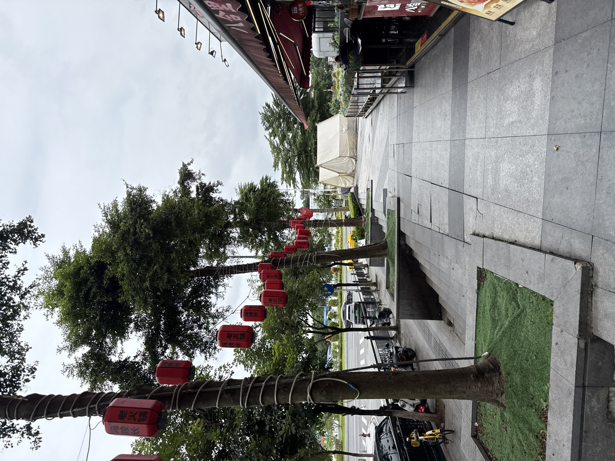 The image depicts a street scene in Haizhu District, China, under an overcast sky. In the foreground, a paved sidewalk runs alongside a row of trees adorned with red lanterns. The trees are wrapped with white rope. To the left of the sidewalk, a street with moving traffic and parked motorcycles is visible. A yellow bicycle is parked on the sidewalk. Further down the sidewalk, past the trees, there are two white tents pitched. On the right side of the image, a building with a red awning and outdoor seating is situated. The building appears to be a restaurant, with signage visible. A person is seated at one of the tables. The overall environment is urban, with a mixture of natural elements like trees and artificial structures such as buildings and tents.