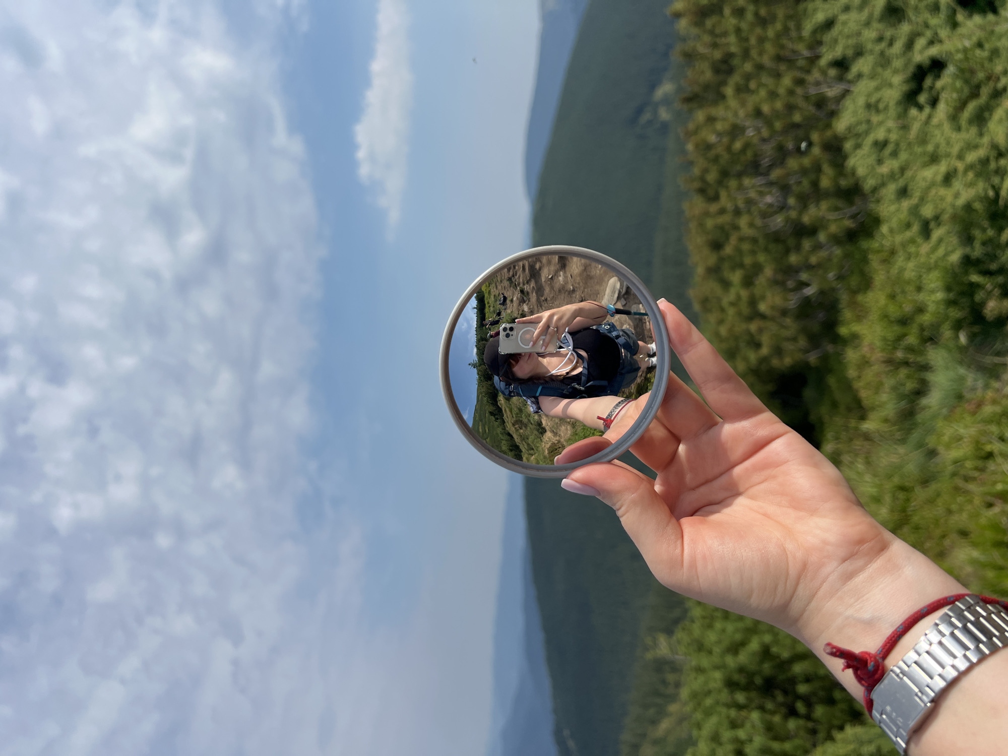 A woman's hand holds a round mirror, reflecting her image as she takes a selfie with her phone. She is wearing a black tank top, grey shorts, and a black baseball cap. A blue backpack is slung over her shoulders, and she holds trekking poles. In the mirror, other hikers can be seen in the background on a mountain trail. The environment is mountainous, with green trees and rolling hills under a blue sky with scattered clouds. The location is stated to be Vorokhta, Ukraine. The woman has a silver watch on her left wrist, and a red string bracelet.