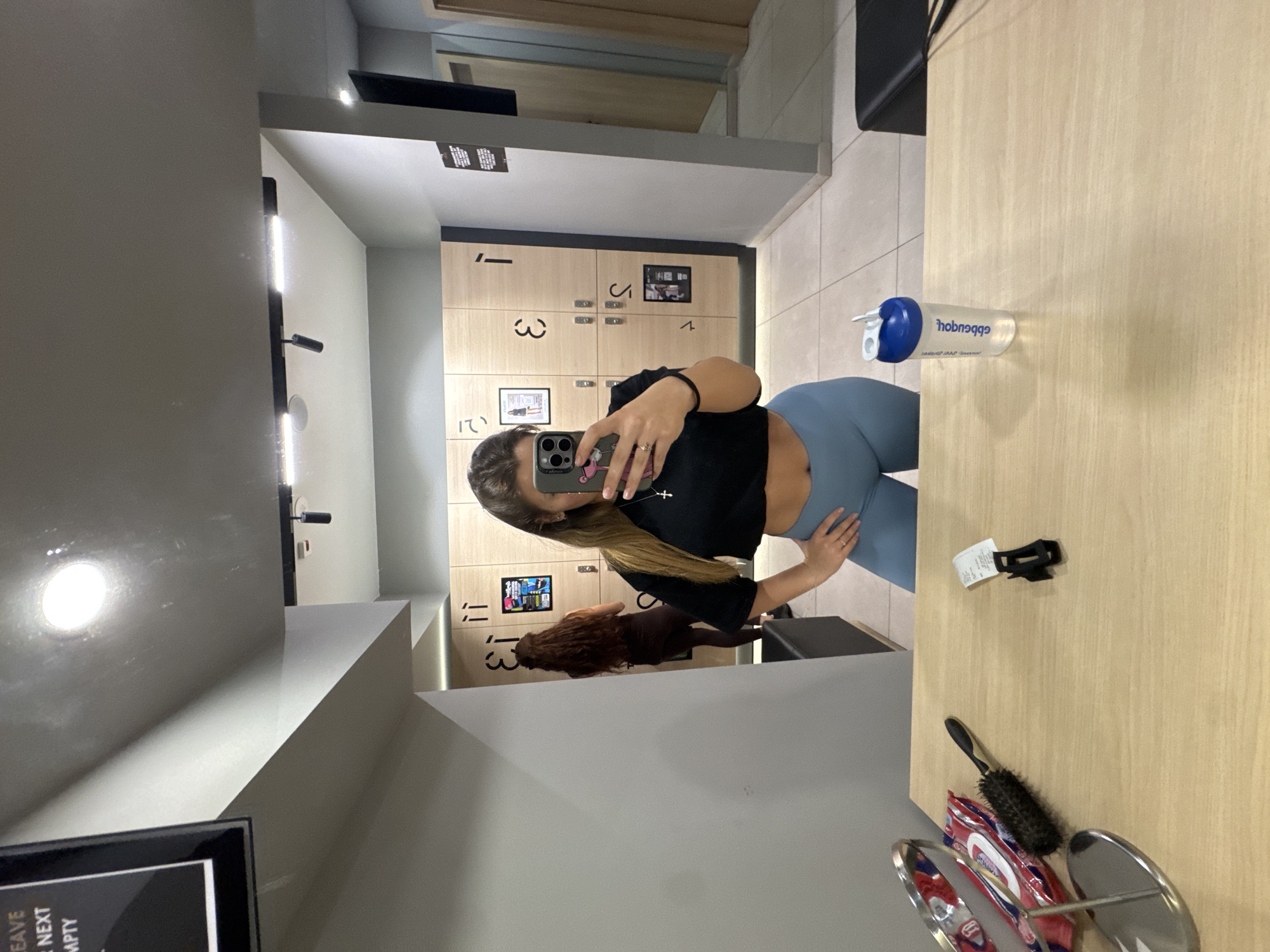 The image captures a mirror selfie taken by a person in what appears to be a changing room or locker room, likely at a gym or fitness facility, given the context of Sofia, Bulgaria. The main subject is a woman with long, light brown hair, wearing a black crop top, light blue leggings, and a silver cross necklace. She is holding an iPhone with a cartoon character phone case to take the photo, and a ring is visible on her finger. Her hand is placed on her hip.
In the background, visible through the mirror's reflection, are wooden lockers with black numbers, including '1', '3', '4', '11', and '12'. Some lockers have small framed pictures or labels attached. Modern strip lighting illuminates the space.
To the right of the main subject, on a wooden countertop or bench, various objects are present: a clear water bottle with a blue cap, featuring the reversed text "hobnɔqqə" (likely "apponeh" unreversed), a black round hairbrush, a black strap or clip, and a red and white packaged item (possibly wipes or a snack), alongside a metallic object that might be a hair tie holder.
A reflection of another person with long, dark, curly hair is partially visible behind the main subject in the lower part of the mirror. A framed sign in the bottom-left corner of the image, partially obscured, shows the words "LEAVE" and "NEXT EMPTY". The overall setting suggests an indoor, well-lit environment.