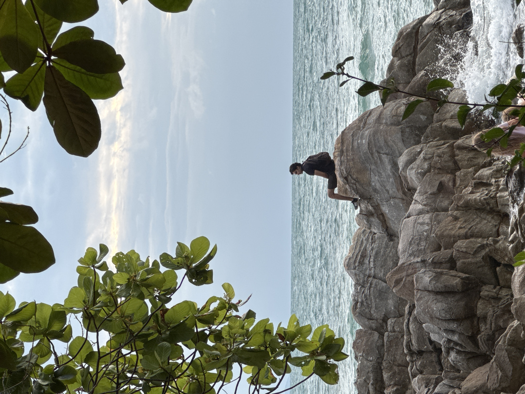 A man is sitting on a large rock by the sea in Ko Pha-ngan District, Thailand. The man is wearing a black t-shirt, black shorts, and a black backpack. He is facing left, looking out at the ocean. The sea is rough, with waves crashing against the rocks. There are trees with green leaves in the foreground, framing the scene. The sky is cloudy and overcast. There is another person, a woman, partially visible sitting on the rocks below.