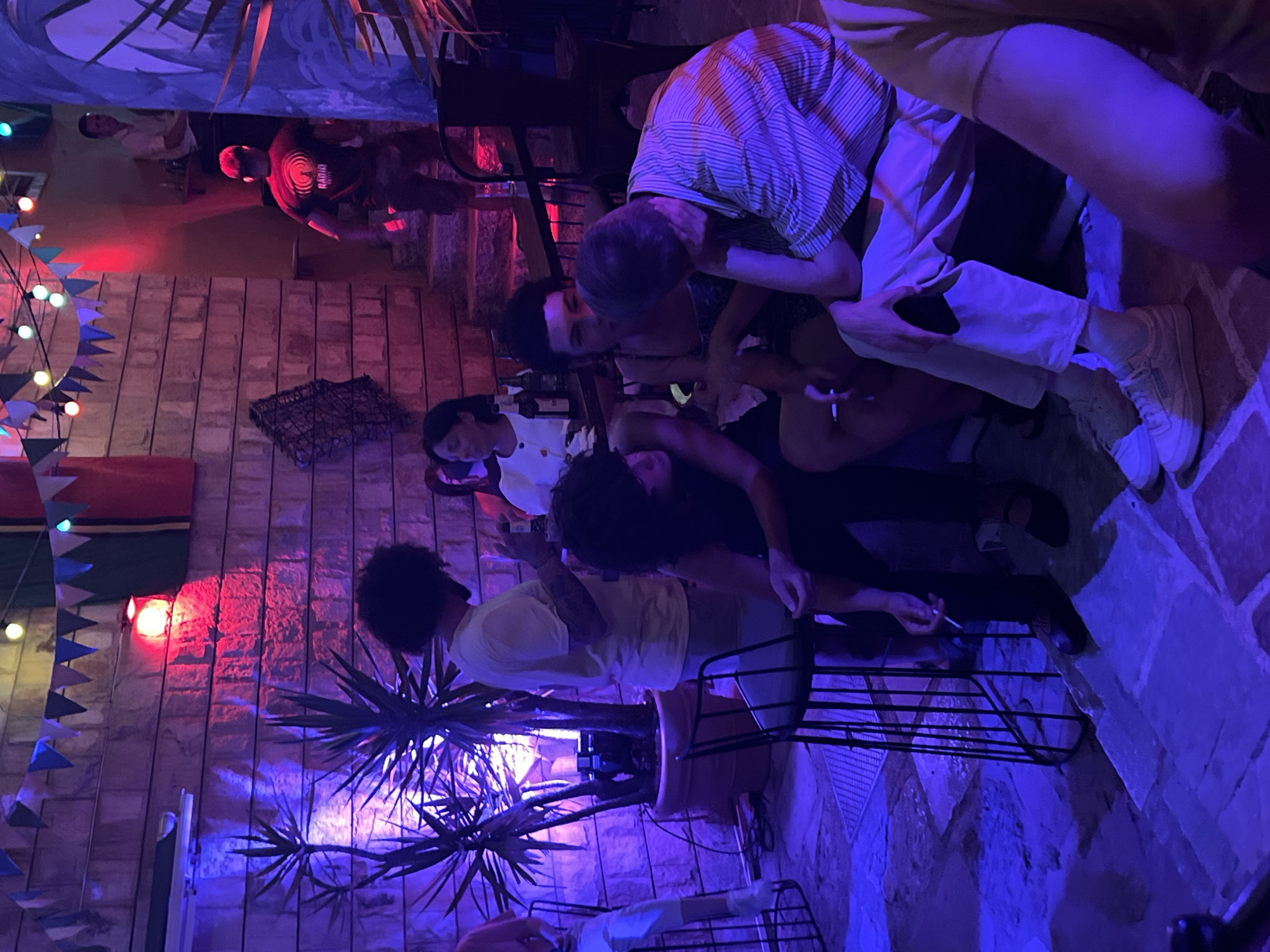 The image depicts an outdoor seating area at night, likely a bar or restaurant, illuminated by various colored lights, predominantly blue and red, and strung fairy lights. In the background, a man wearing a black t-shirt with a circular logo and the word "RADIO" is descending a stone staircase. Above, colorful triangular flags are strung across the top of the scene. In the mid-ground, a woman in a white top sits at a table with bottles. To the left, several people are seated on the ground and on stools, some interacting and one person holding a cigarette. A large potted plant with spiky leaves stands to the left, casting shadows. The ground is paved with irregular stones. The overall atmosphere suggests a casual, social gathering in a dimly lit, atmospheric setting, possibly in Herceg Novi, Montenegro, given the context.