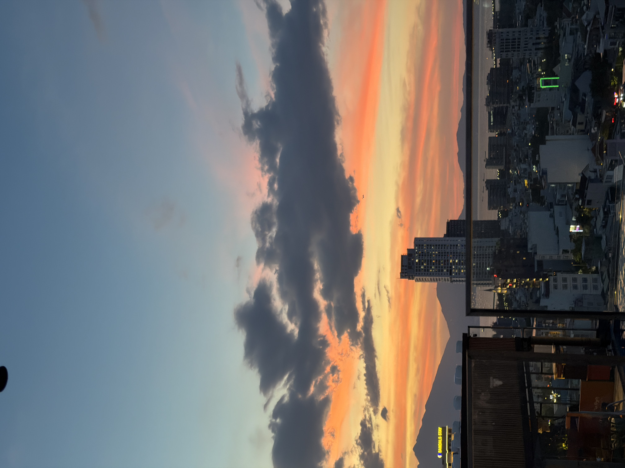 This is a vertical, eye-level, long shot of the city of Da Nang, Vietnam, during a sunset. The sky is painted with a gradient of pale blue at the top, transitioning to vibrant oranges and pinks as it meets the horizon. Dark, heavy clouds are scattered across the sky, some tinged with orange light from the setting sun. In the foreground, there are parts of buildings and balconies, suggesting this photo was taken from a rooftop or high vantage point in a building. In the mid-ground, a city skyline is visible with numerous buildings of varying heights. Notably, a tall, modern skyscraper stands out, and in the distance, mountains can be seen. The sea is also visible on the right side of the frame. The overall impression is of a bustling city at dusk, bathed in the dramatic colors of a sunset. There is a small object, possibly a drone, in the upper portion of the sky.