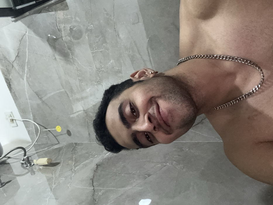 A selfie shows a man with dark hair and a slight beard, wearing a silver chain. He is shirtless and smiles slightly at the camera. The background consists of gray marble wall tiles and some electrical cords, a switch and a yellow tag on the wall. The image is oriented sideways, with the subject's head tilted to the left.