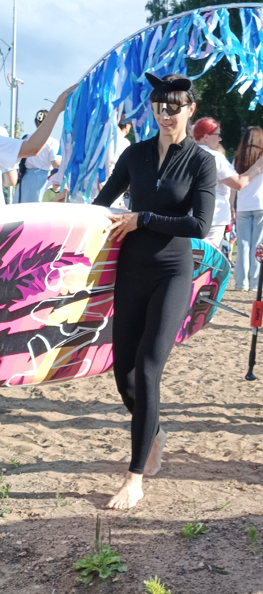 An outdoor, daytime, eye-level, full-body shot shows a woman walking barefoot on a sandy beach, holding a colorful surfboard. The woman is wearing a black, long-sleeved, full-body suit and black sunglasses. She has dark hair and appears to be smiling. Behind her, a decorative archway is adorned with blue, white, and purple streamers. Other people are visible in the background, some wearing white shirts and light-colored bottoms, and one person with red hair. The environment is a beach with sand and sparse vegetation, under a bright sky.