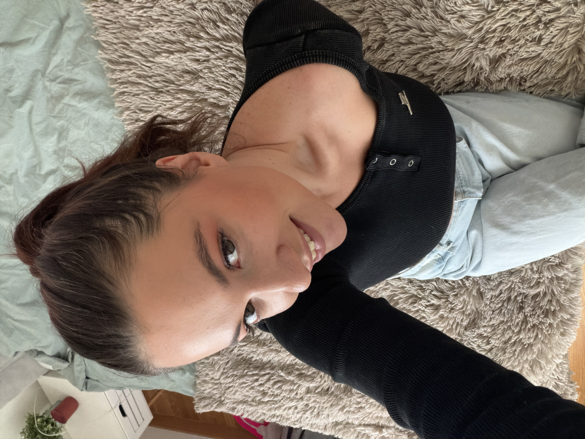 A woman lies on her back, positioned on a shag rug that transitions into a light blue bedsheet on the left side of the image. The rug is light brown. The woman is wearing a fitted, long-sleeved black top that features a button-down detail and a metallic pin near the neckline. She is also wearing light blue denim jeans. Her hair is pulled back into a bun. Her face is turned slightly towards the camera, and she is smiling with visible teeth. To the lower left corner of the image, parts of white furniture, including a small nightstand and what appears to be a houseplant in a red pot, are visible. Given the indoor context, the location is presumably in an apartment or home. The image provides no information to confirm the location is Belgrade, Serbia.