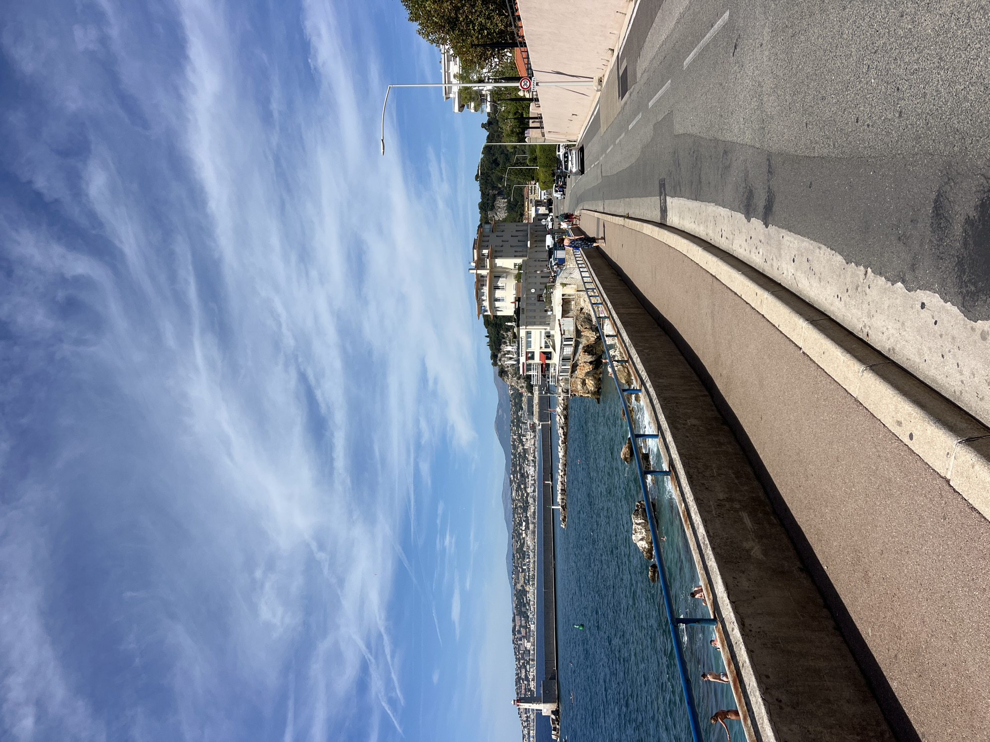 This image captures a picturesque scene along the coast of Nice, France, likely on the Promenade des Anglais or a similar seaside road. The foreground features a paved walkway next to a concrete barrier overlooking the deep blue Mediterranean Sea. Beyond the barrier, a few individuals are visible near the water's edge, some appearing to be sunbathing or wading.
The sea stretches out to a long breakwater with a lighthouse at its far end, and a city skyline with numerous buildings and hills in the background. A few small boats are scattered in the water.
To the right of the walkway, a road curves away from the viewer. Several cars are driving on the road, and a traffic sign is visible on a pole. Buildings line the right side of the road, some with balconies and awnings, suggesting a mix of residential and commercial structures. Trees and vegetation are also present.
The sky above is a vibrant blue, dotted with wispy white clouds that create a dynamic texture. The overall atmosphere is one of a sunny, pleasant day in a coastal city. The scene is captured from a slightly elevated perspective, looking down the road and out towards the sea.