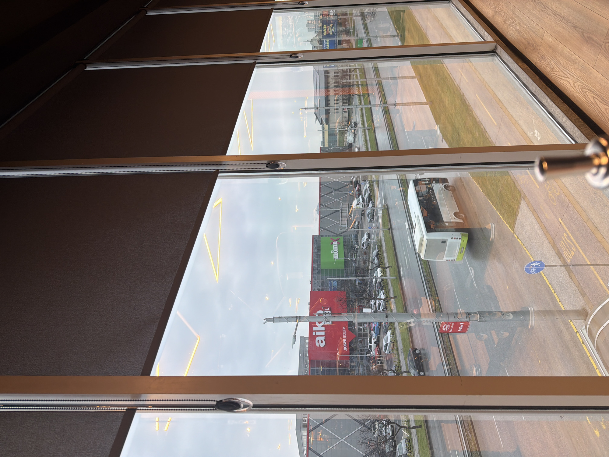 The image shows a view from inside a building, looking out through a multi-pane window. The window panes have partial, dark brown blinds covering the top portions. The view outside reveals a street scene, which includes buildings, cars, a bus, and roadside elements. The street appears wet, possibly from rain. The most prominent buildings visible are a red building with the word "AIK" written on it in large white letters and a green building with "Tromp" written on it. There is a bus in the street, and other cars can be seen. There is signage and poles along the roadway. The photograph is taken from an elevated position, looking down at the street below. The location is Plovdiv, Bulgaria, based on the "AIK" advertisement.