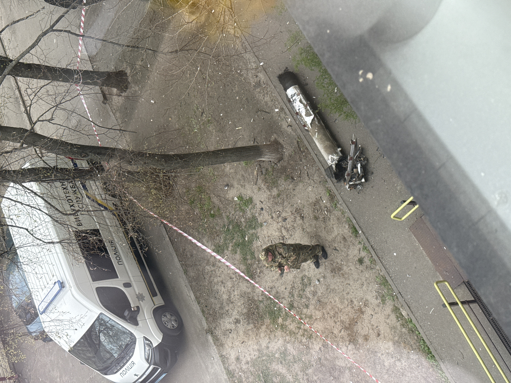 This image depicts a scene in Kyiv, Ukraine, showing the aftermath of a strike. Visible elements include a damaged missile or rocket component lying near a sidewalk, a police vehicle with Ukrainian lettering, and a uniformed individual inspecting the debris. Red and white tape cordons off the area. Trees with bare branches and green grass are also visible, suggesting it is either late winter or early spring. The image appears to be taken from an elevated position.