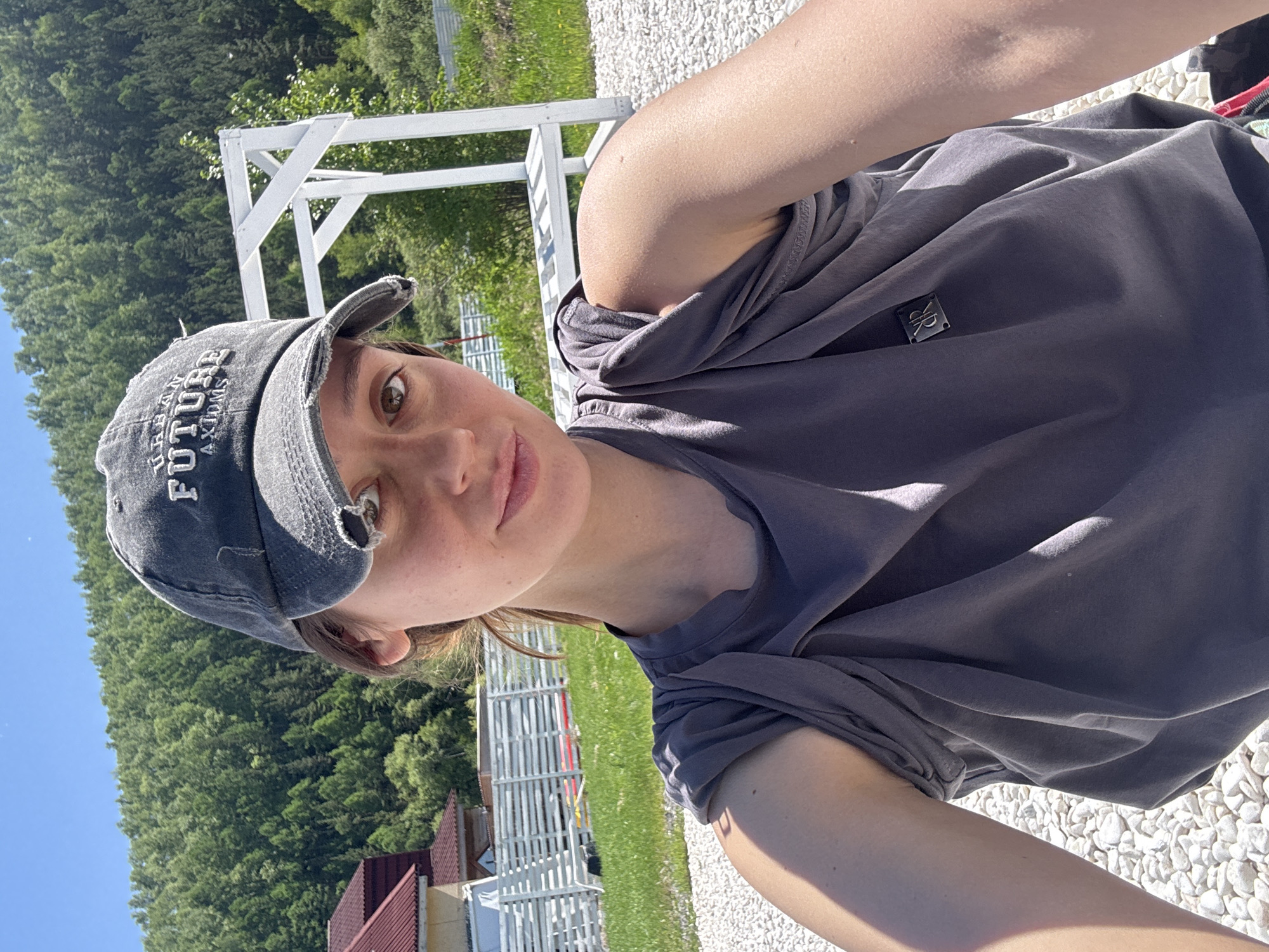 A selfie-style portrait captures a young woman outdoors in what appears to be a sunny, landscaped area in Дрокино, Russia. The image is rotated 90 degrees counter-clockwise, showing her face in the upper central portion.
The subject is a young woman with light brown hair and brown eyes, looking directly at the camera with a neutral to slightly contemplative expression. She wears a distressed dark grey baseball cap with white embroidered text, which reads "URBAN FUTURE AXIO.S" across the front. Her attire includes a plain grey short-sleeved t-shirt. A small rectangular black emblem with silver "VK" or "VR" initials is visible on the left side of her t-shirt, near the shoulder.
The background reveals a bright, clear blue sky and lush green forested hills under strong sunlight, suggesting a clear day. In the middle ground, there are white wooden structures, possibly part of a pergola or fence, and further back, the roof of a building with red tiles. The ground beneath her is covered in small, light-colored pebbles or gravel, adding to the bright, well-maintained outdoor setting. The strong sunlight casts some shadows, highlighting the textures of her clothing and the outdoor environment.