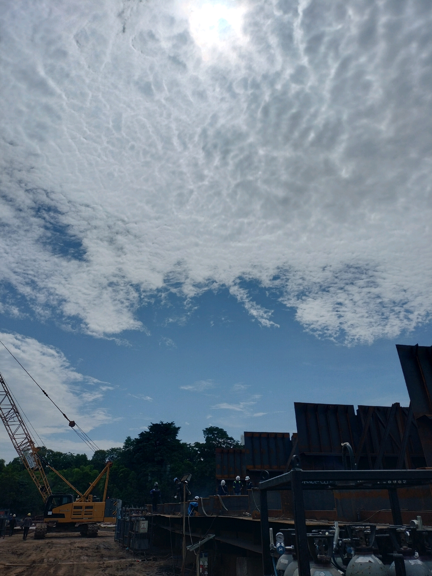 In Batam, Indonesia, a construction site is depicted with a large yellow crane on the left side of the frame. The crane is positioned on a muddy ground, with a large structure made of steel panels in the background. Several workers in hard hats and safety vests are visible on the steel structure, some appearing to be welding. The sky above is filled with a dramatic cloud formation, with patches of blue sky visible. Trees line the background, suggesting an outdoor, industrial setting. The overall scene conveys an active construction project underway.