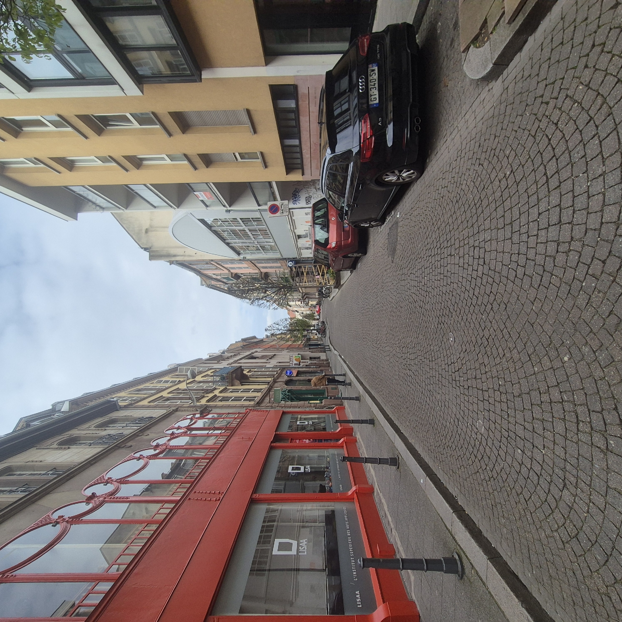 Here is a detailed description of the image:
**Overview:**
The image depicts a street scene in Strasbourg, France. The camera's perspective is slightly tilted, capturing the street's architecture and details.
**Setting and Architecture:**
The street is paved with cobblestones and lined with buildings on both sides. The buildings exhibit a mix of architectural styles. On the left side of the street, there's a distinctive red building with large glass windows, which appears to be the "LISAA" - L'Institut Supérieur des Arts Appliqués. The windows of LISAA have a red, decorative framework. Other buildings are visible with yellow and grey facades.
**Vehicles:**
Parked along the right side of the street are several cars. Most notably there is a black Audi A3.
**People:**
In the middle of the shot, it looks like one or two people are walking down the street.
**Details:**
Black metal bollards are positioned along the edge of the sidewalk near the LISAA building, serving as a barrier. The sky is overcast with a pale blue hue. Trees are seen sporadically along the street. Street signs and other urban elements are also visible.
**Location:**
The image shows a street in Strasbourg, France, likely near the LISAA higher learning institution, as prominently displayed on the building.