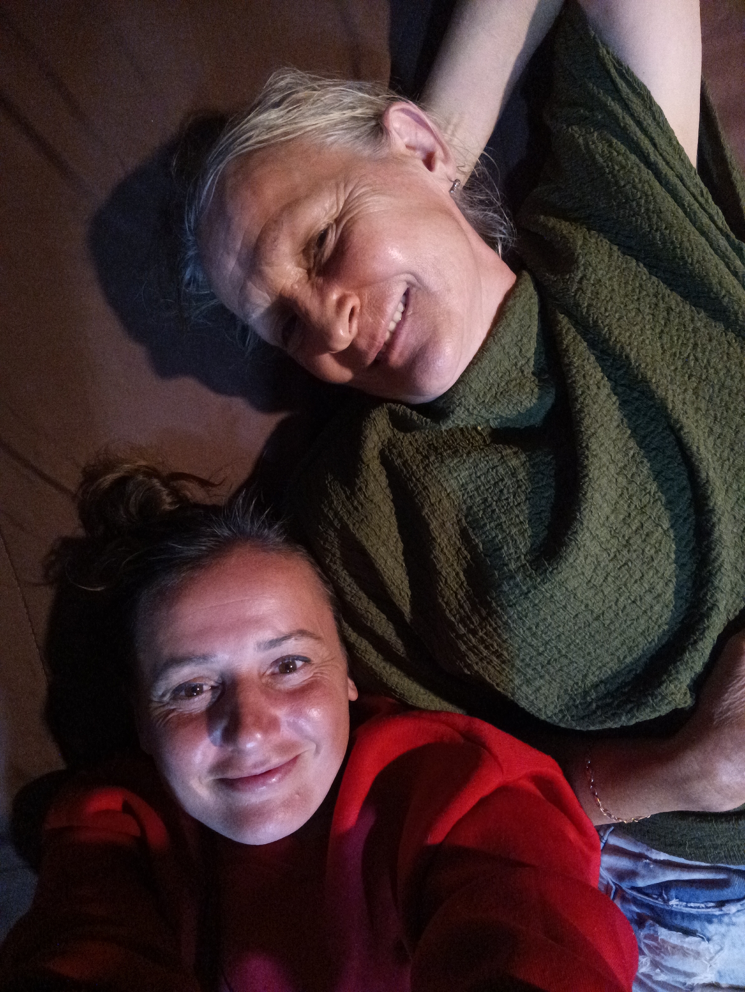 Two women lie on a brown surface, likely a floor or bed, in Ban Tak Daet, Thailand. The woman on the bottom wears a red shirt and has her hair in a bun. The woman above her wears a green, textured garment. Both women are smiling and appear to be looking at the camera, suggesting a selfie. An anklet is visible on the arm or leg of the woman in green. The lighting is uneven, with shadows obscuring some details.
