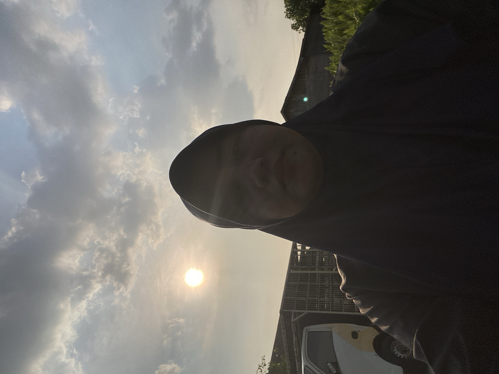 A selfie is taken by a person wearing a black hijab outdoors, with the sun visible in the upper left quadrant of the frame. The background includes a white and yellow truck on the left, a weathered wooden structure with metal fencing behind it, and a small building with a dark roof and wooden siding on the right. A row of green plants is visible in the foreground on the right. The sky is filled with clouds, illuminated by the sun. The scene appears to be in a rural or semi-rural area, possibly on the outskirts of Banjarmasin, Indonesia, given the presence of the truck and the type of structures. The lighting suggests it is late afternoon or early evening.