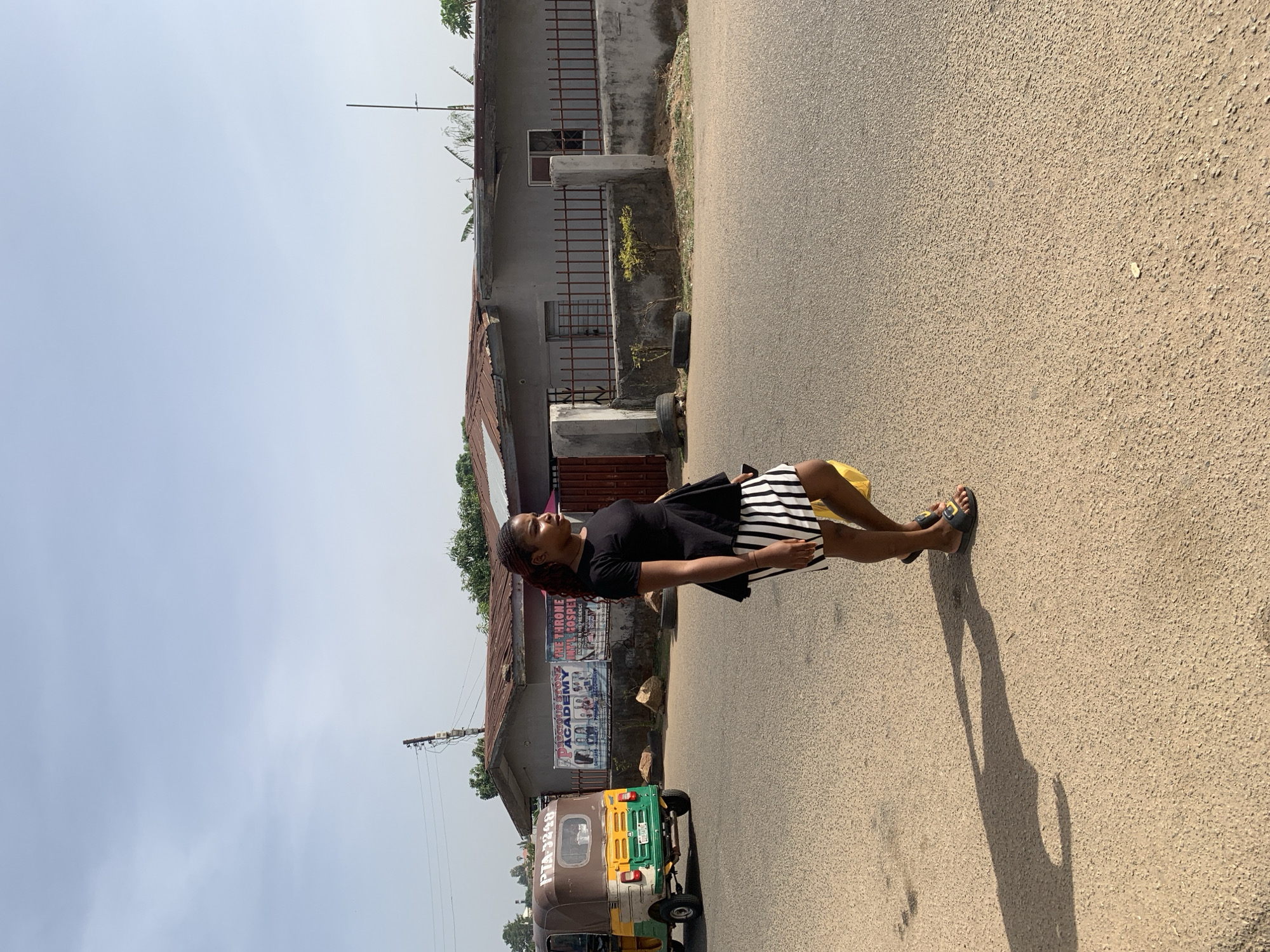 The image captures a street scene, likely in Jos, Nigeria, featuring a woman standing on the side of a paved road. The woman, positioned towards the right of the frame, is seen in profile, looking slightly upwards. She is wearing a black short-sleeved top and a black and white striped skirt, with dark sandals on her feet. A yellow object is visible near her waist. Her long shadow stretches across the road, indicating the sun is low in the sky, suggesting either early morning or late afternoon.
In the background, a three-wheeled auto-rickshaw, commonly known as a Keke Napep, is partially visible, displaying brown, yellow, and green colors. The license plate or identification number "PIA-3240" is visible on its side. Behind the vehicle and the woman are several single-story buildings constructed with concrete and topped with corrugated iron roofs. One building features banners or posters with visible text including "OUR HOPE ACADEMY" and "THE THRONE ROYAL GOSPEL." The sky above is a bright, pale blue, suggesting clear and fair weather conditions. The scene depicts a slice of daily life on a sunny street.