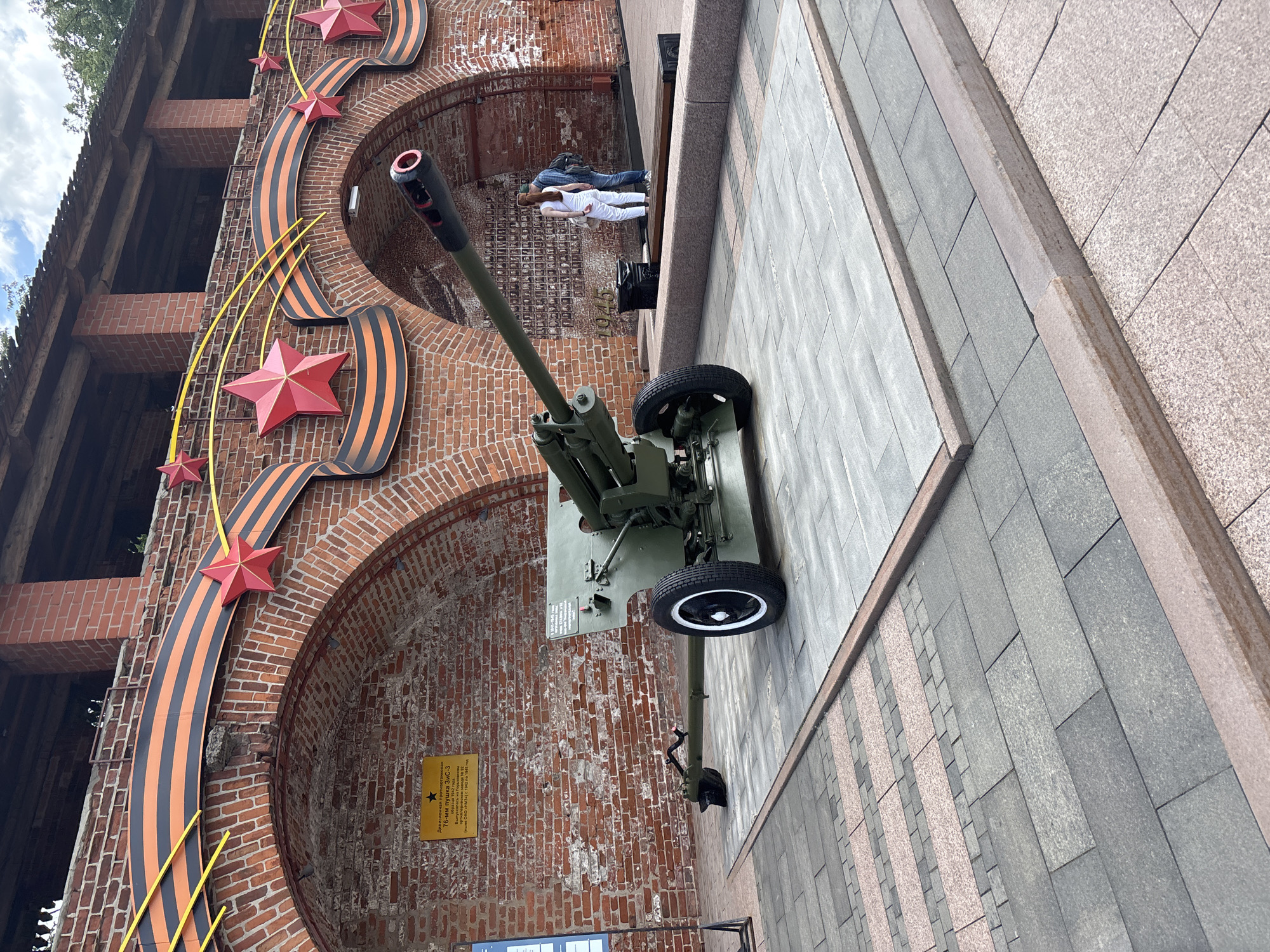 This outdoor image, taken in Nizhny Novgorod, Russia, features a static display of military equipment and commemorative decorations next to a brick building. In the foreground, a green 76-mm ZiS-3 cannon, dating from 1942, is prominently displayed, facing left on a paved ground surface. Its wheels and barrel are clearly visible.
The cannon is situated in front of a multi-story brick building with distinct arches, suggesting historical or institutional architecture. Adorning the brick wall are large decorative elements, including multiple red stars, yellow swooping lines, and a prominent black and orange striped ribbon, emblematic of the Russian Ribbon of Saint George, often associated with military valor, particularly from World War II.
In the mid-ground, under one of the arches of the brick building, two people are visible. One, dressed in white, is sitting or lying down, while the other, wearing a backpack, is standing nearby. They appear to be resting or observing the surroundings.
A yellow plaque on the brick wall next to the cannon provides details in Cyrillic: "Дивизионная противотанковая 76-мм пушка ЗиС-3 образца 1942 года. Выпущена на производственных мощностях Сталинградского Завода №92 им. И. В. Сталина (КБВН) в период с 1942 по 1945 год." This translates to: "Divisional anti-tank 76-mm cannon ZiS-3, model of 1942. Produced at the production capacities of Stalingrad Factory No. 92 named after I.V. Stalin (KBVN) in the period from 1942 to 1945."
The scene is set during the daytime, under an overcast sky, as suggested by the soft, diffused lighting. The overall impression is that of a historical military memorial or museum exhibit, honoring the Great Patriotic War.