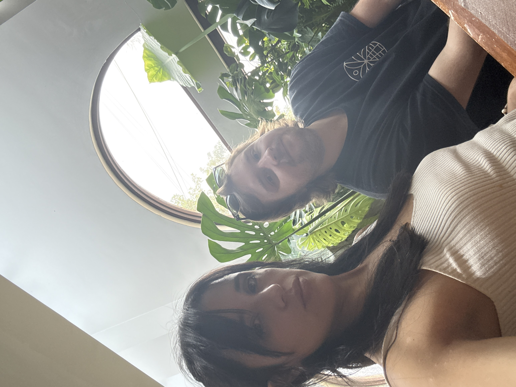 The image shows two individuals indoors. A woman with dark hair is in the foreground wearing a light-colored ribbed top. Behind her is a man with long hair and a beard, wearing a dark t-shirt with a graphic design. They are positioned in front of a window, with lush green plants filling the background. The window is set into a light-colored wall. The perspective suggests the photo was taken from a low angle looking upwards. The location may be in Cuautitlán Izcalli, Mexico.