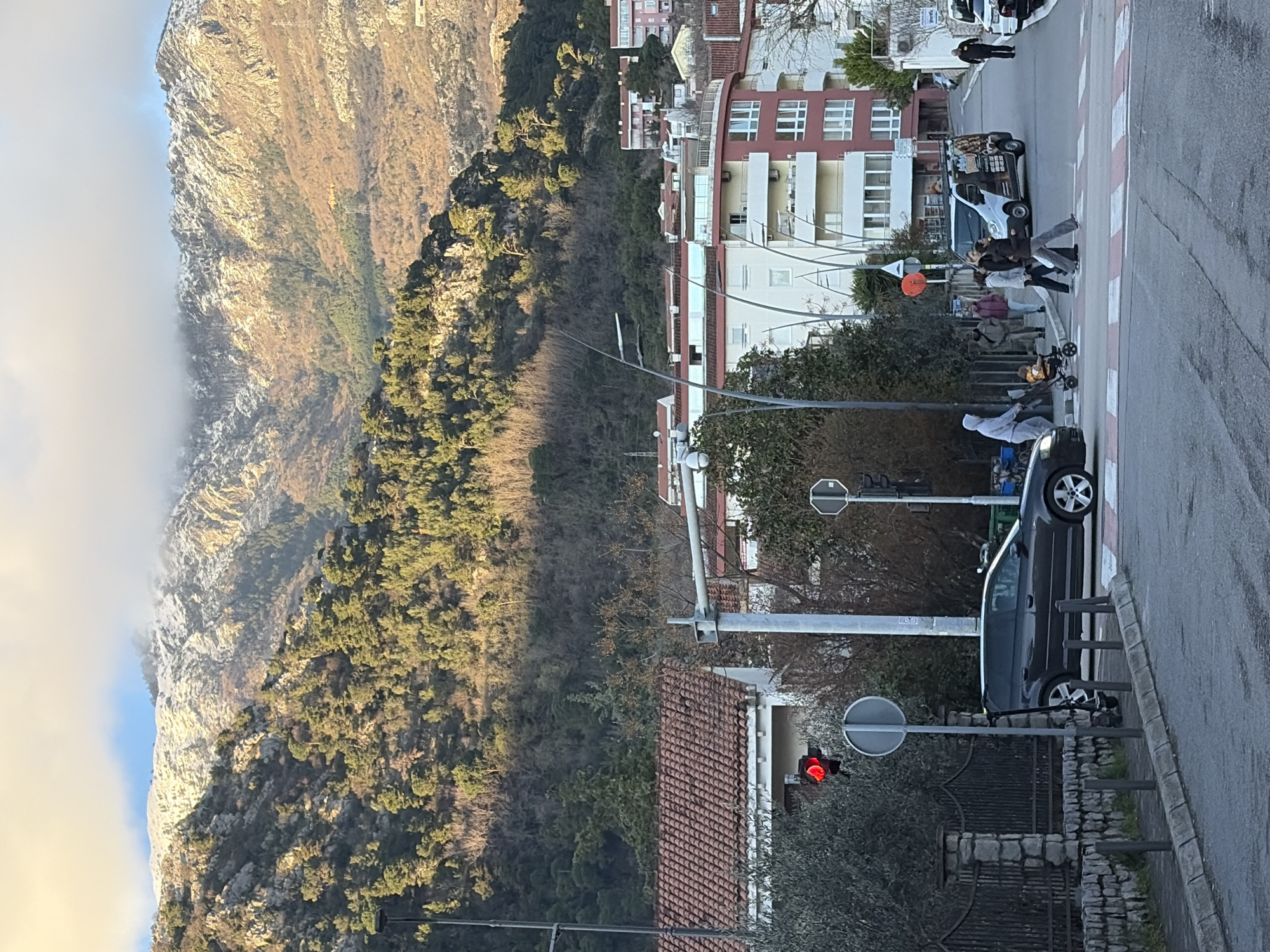 This image depicts a street scene in Budva, Montenegro, with a prominent mountainous backdrop.
**Location:** The scene is an urban intersection in Budva, Montenegro, with residential buildings and a street leading uphill.
**Environment:**
The background features large, rugged mountains partially covered with snow on their upper slopes, with some cloud cover near the peaks. Below the snowline, the mountainsides are heavily forested with a mix of evergreen and deciduous trees, displaying varied shades of green and brown.
The foreground and midground show an asphalt road with a marked pedestrian zebra crossing. A sidewalk runs along the left side, bordered by a low stone wall with a black metal fence and small bollards. Street infrastructure includes light poles with modern fixtures, a traffic light displaying red on the left side of the intersection, and various road signs.
**Objects:**
A dark grey hatchback vehicle is stopped at the pedestrian crossing, facing right. An electric scooter is leaning against a signpost on the sidewalk near the hatchback. Further up the road to the right, a white commercial van with graphic decals on its side is visible. Additional vehicles are parked or driving further uphill.
Buildings are primarily multi-story residential structures, some featuring red-tiled roofs and white facades, while others incorporate reddish-brown sections and balconies. Lush green and bare deciduous trees and shrubs are interspersed throughout the urban landscape.
**People and Actions:**
A group of five pedestrians is actively crossing the street on the zebra crossing from left to right.
1. A person wearing a light grey hooded sweatshirt is pushing a baby stroller containing a small child dressed in an orange top.
2. Immediately behind them, three adults are walking together. One individual wears a dark jacket, another a light-colored jacket, and a third is partially obscured.
3. Further up the street on the right side, a lone male individual in a dark jacket is walking away from the camera.