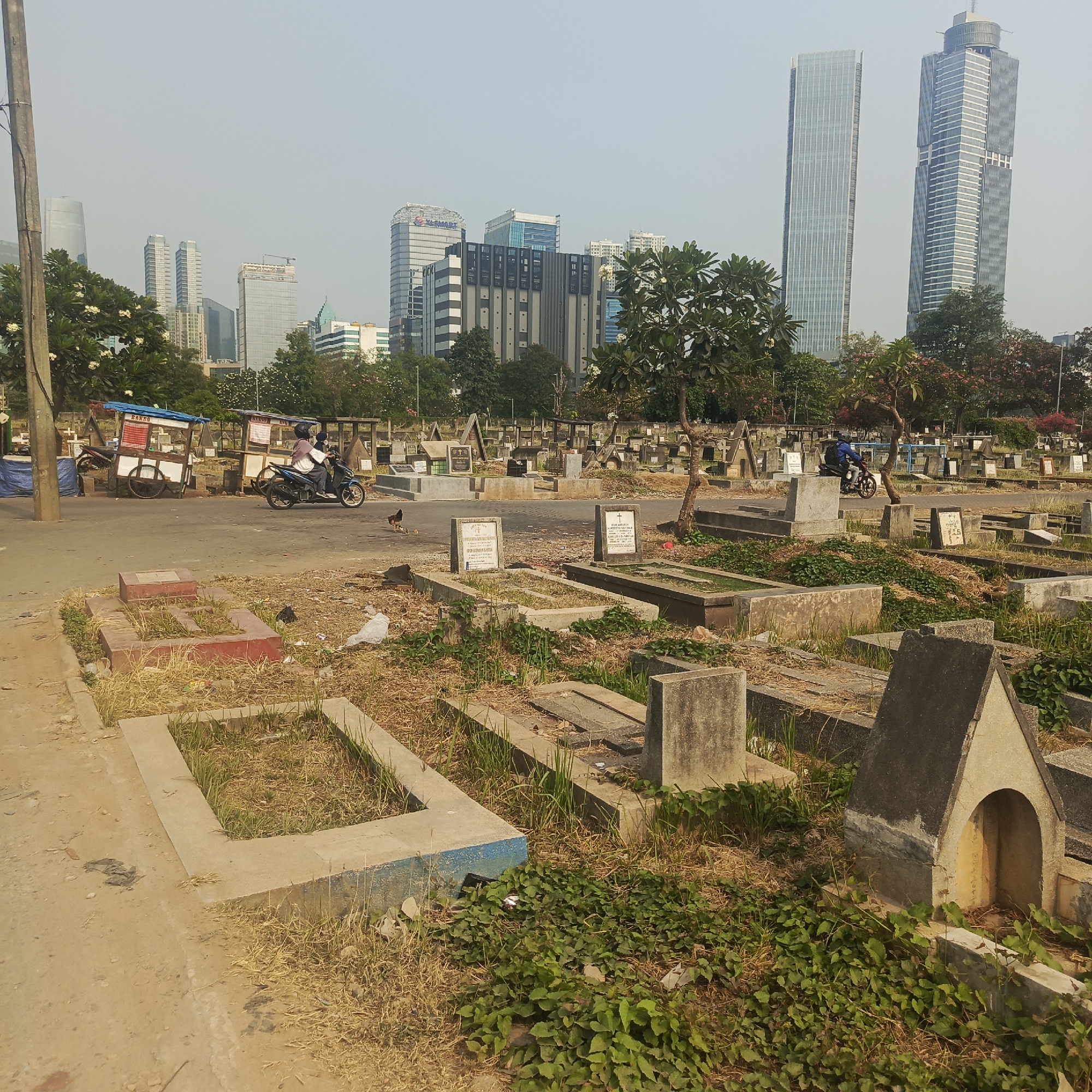 The image depicts a cemetery in South Jakarta, Indonesia, with modern skyscrapers visible in the background. In the foreground, numerous graves and tombstones are scattered across the landscape, many appearing weathered and overgrown with grass and vines. A dirt path or road runs through the cemetery, with several food carts visible on the side. A person is riding a motorcycle past one of the food carts, while another motorcycle is parked nearby. A chicken is also visible on the ground near the road. The sky is hazy and overcast, typical of urban air quality in the region. The overall scene presents a stark contrast between the traditional burial grounds and the contemporary urban development.