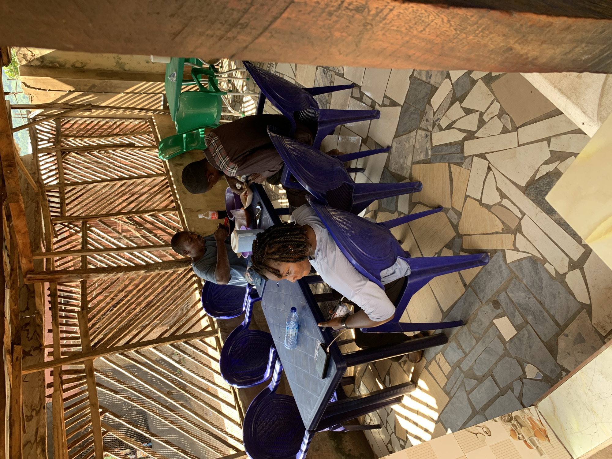 The image depicts an outdoor dining area with a rustic, open-air roof structure made of wood and bamboo. Three individuals are seated at a blue plastic table, engaged in eating and drinking. The table is laden with plates of food, a bottle of Coca-Cola, a water bottle, and a white pitcher. Two men are seated at the far end of the table, while a woman with braided hair is seated in the foreground, looking at her phone. The floor is tiled with irregular pieces of stone and ceramic. In the background, green plastic chairs and tables are visible, suggesting a larger seating area or adjacent establishment. The overall ambiance is casual and informal, likely a local eatery in Lokoja, Nigeria, given the architectural style and the presence of common food and drink items. The lighting suggests it is daytime, with sunlight casting strong shadows through the wooden lattice of the roof.