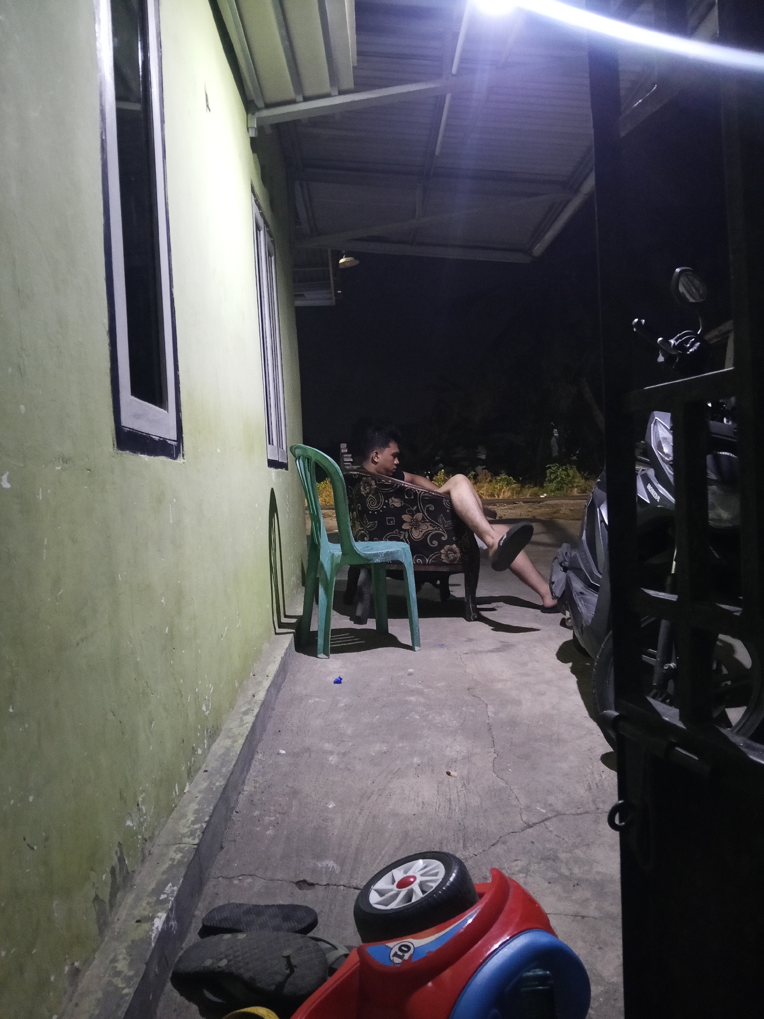 A young man is sitting in a chair on a concrete patio in front of a pale green house at night. He is wearing a dark shirt and shorts, and his legs are extended. A turquoise plastic chair is to his left. To the right of the man, a motorcycle is parked. In the foreground, a red and blue toy car is visible on the concrete. The scene appears to be in an outdoor residential area in Lubuklinggau, Indonesia.