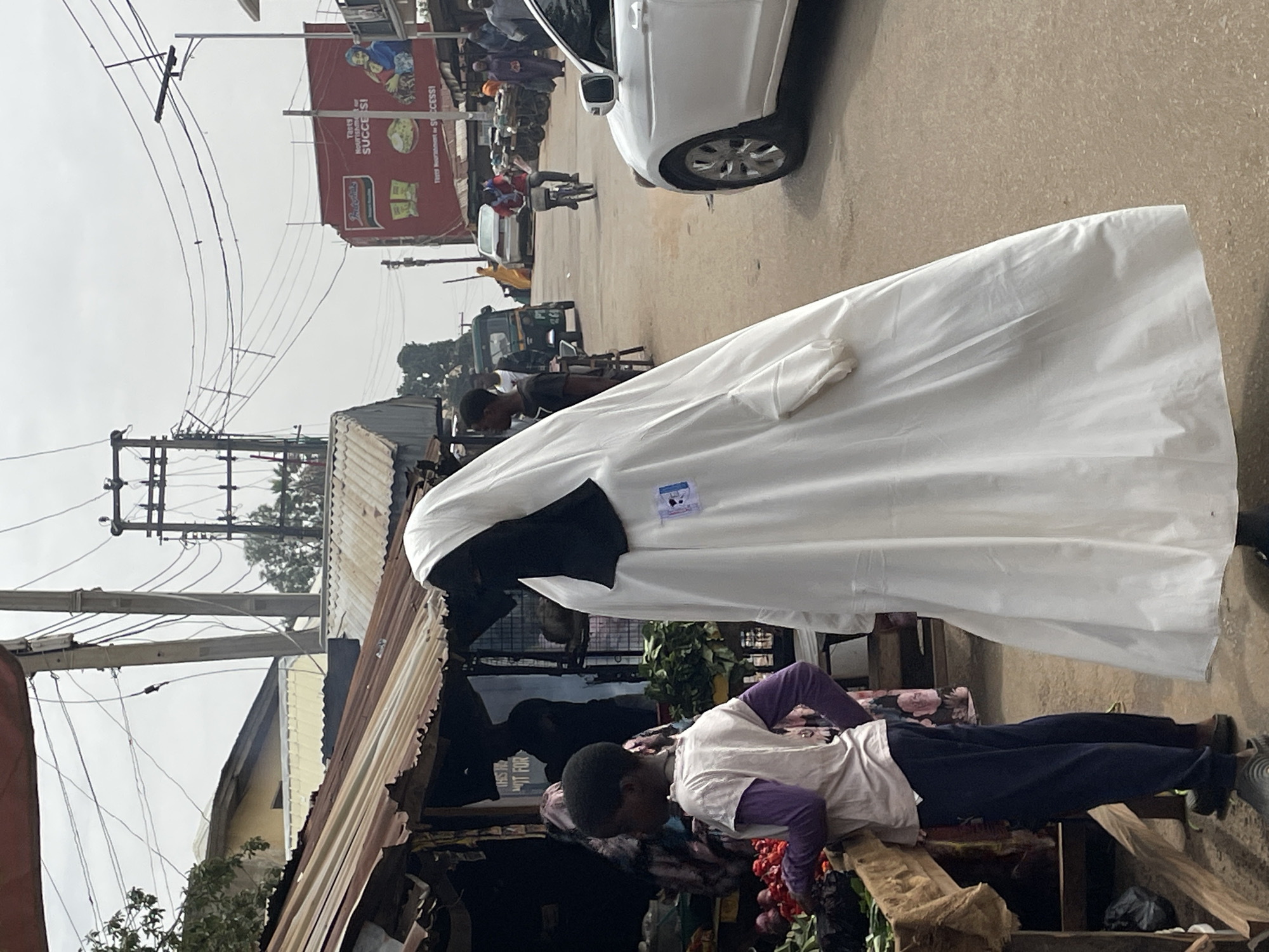 A street scene in Jos, Nigeria. A person in a large white robe with a black head covering stands near a roadside vendor selling produce. Overhead, there are power lines and a billboard. A white car is parked on the street, and other people and vehicles are visible in the background.