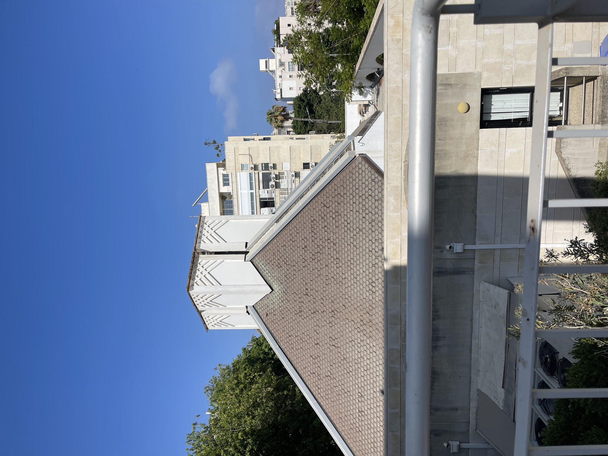 The image captures a view of several buildings and trees under a clear blue sky in Tel Aviv, Israel. In the foreground, a light-colored metal railing runs horizontally across the bottom half of the frame. Behind the railing, a light-colored stone building with a small black-framed window and a yellow circular light fixture is visible. To the left of this building, there is a covered, slanted structure with outdoor air conditioning units visible beneath it.
Dominating the middle ground is the tiled roof of a building with a distinctive white, stepped, decorative structure on top, reminiscent of an Art Deco style. This roof is composed of small, overlapping, curved, brown tiles. To the left of this tiled roof, lush green trees partially obscure the view.
Further in the background, several other buildings are visible. One is a light-colored, multi-story apartment building with numerous windows and air conditioning units. Another building, further to the right, is also light-colored and appears to be taller. A lone palm tree stands between two of the background buildings. The sky is a vibrant, cloudless blue. The overall impression is of an urban environment bathed in sunlight.