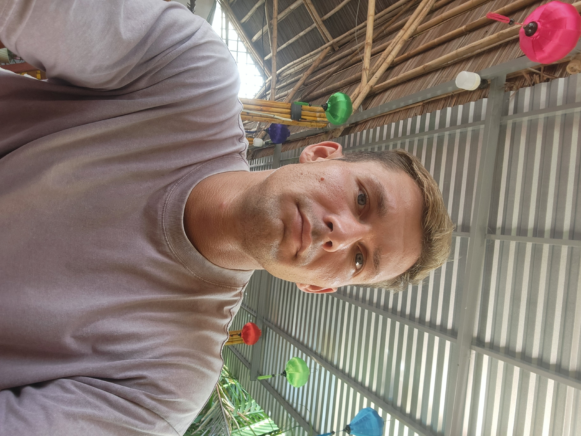 A man with blonde hair is taking a selfie under a covered patio. He is wearing a grey t-shirt. In the background, there is a thatched roof and corrugated metal roof. There are several colorful lanterns hanging from the ceiling, including pink, green, blue, and red. There are also bamboo poles and palm fronds visible. The man's face is the main focus of the image. He has fair skin and light blue eyes. The lighting suggests it is daytime, and the overall scene appears to be a relaxed, outdoor setting, possibly a cafe or market in Da Nang, Vietnam, given the presence of lanterns and tropical foliage.